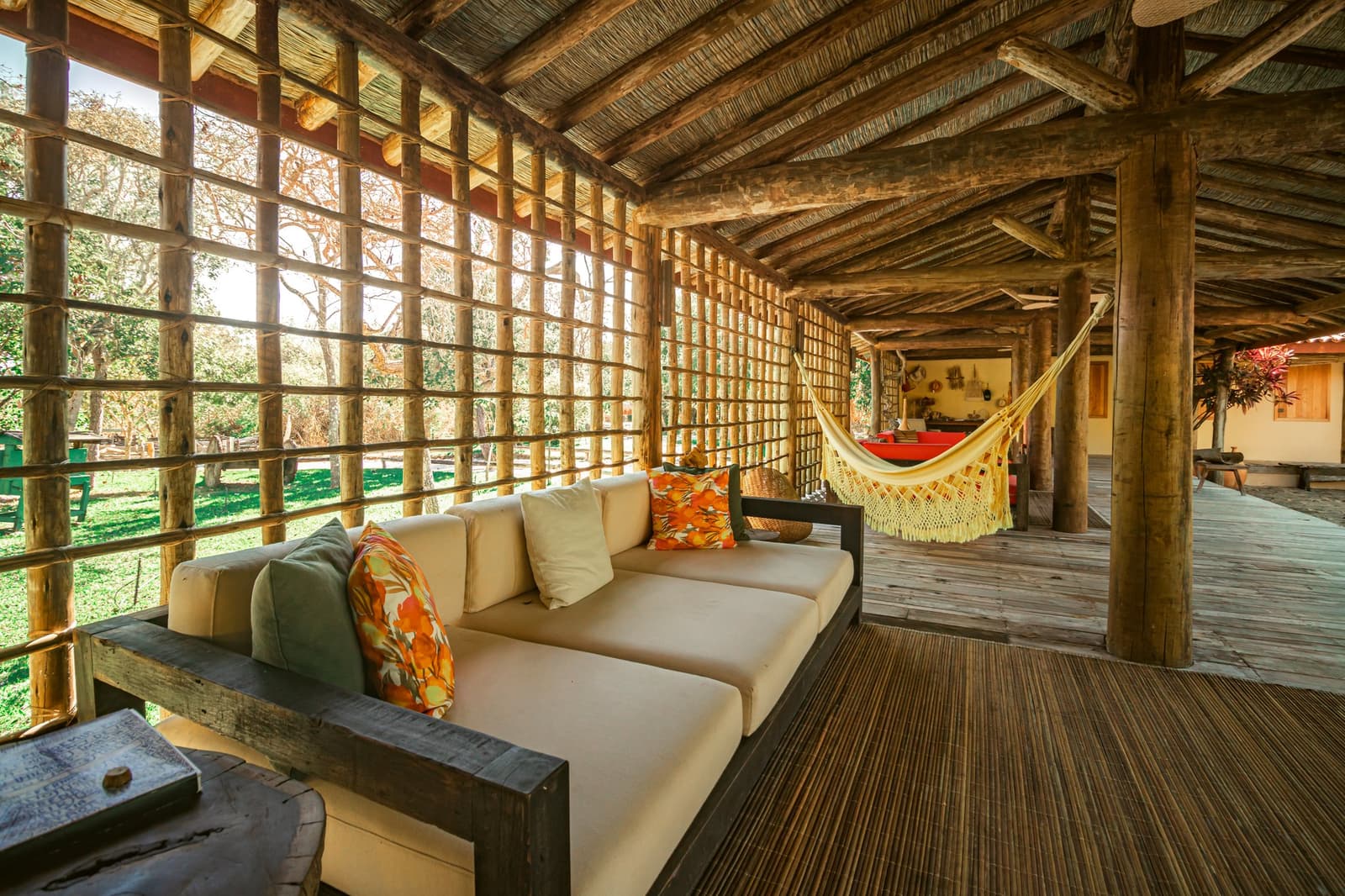 The kind of porch that makes you forget you had plans. Open-air veranda at Pousada Trijunção with a cream sofa, colorful cushions, a fringed hammock, and a rustic log-and-thatch structure overlooking lush Cerrado gardens