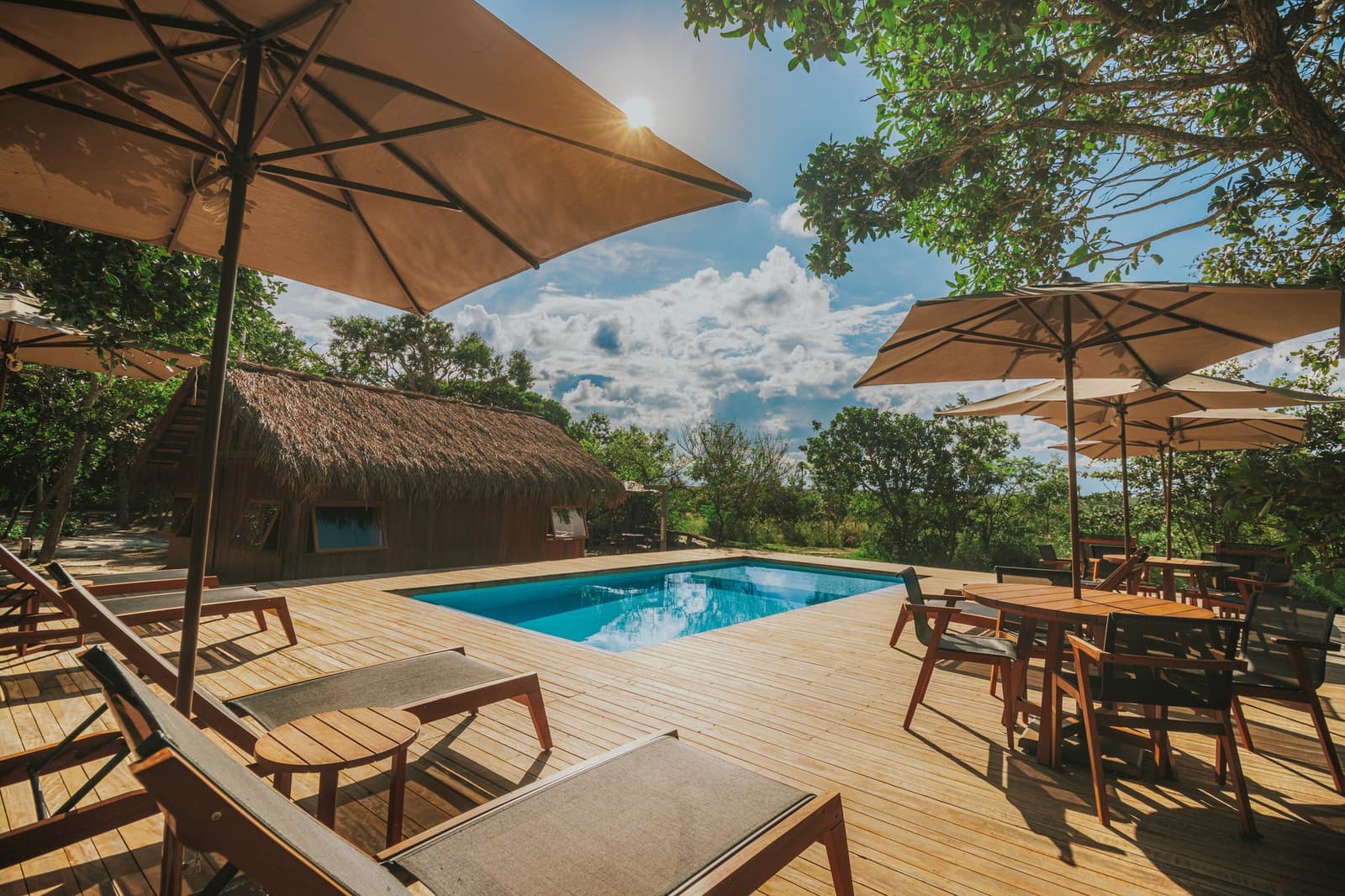 Cold water, warm wood, and the cerrado humming just beyond the fence. Sun-drenched wooden pool deck at Pousada Trijunção with lounge chairs, canvas umbrellas, a thatched-roof building, and green Cerrado vegetation under a blue sky