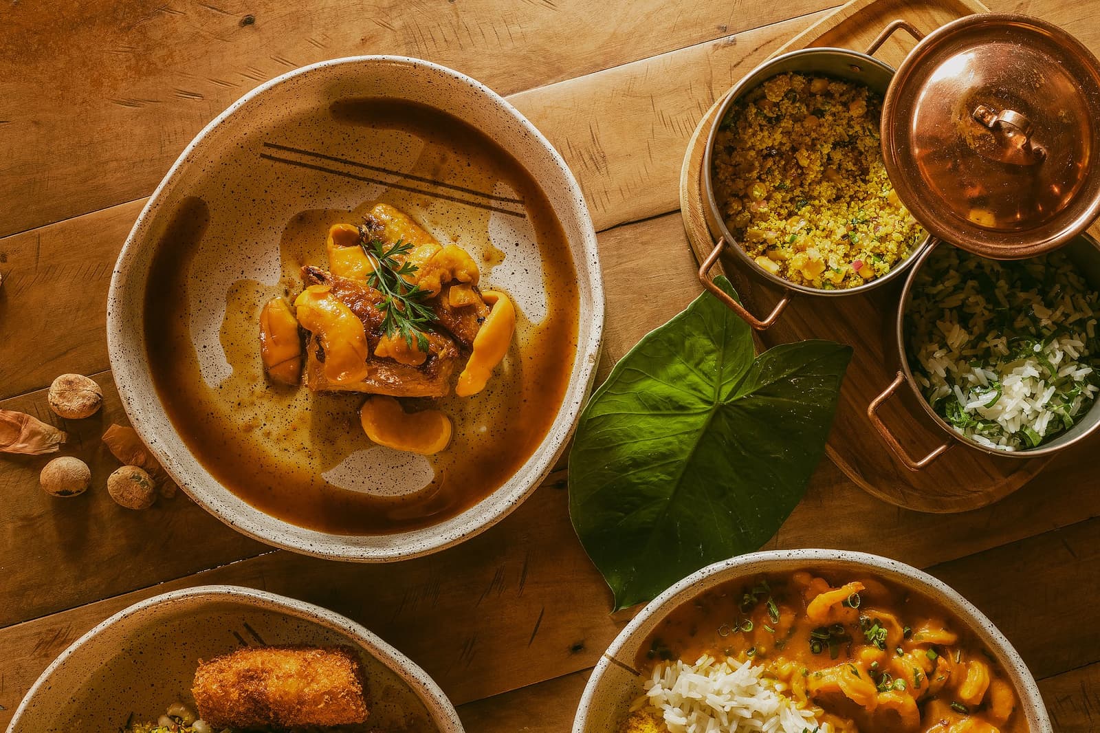 The kind of lunch that makes you forget you had plans for the afternoon. Overhead view of a Pousada Trijunção meal spread featuring a rich braised meat stew in a speckled ceramic bowl, golden farofa and herb rice in copper pots, and a creamy seafood stew with white rice, all arranged on a rustic wooden table with a tropical leaf