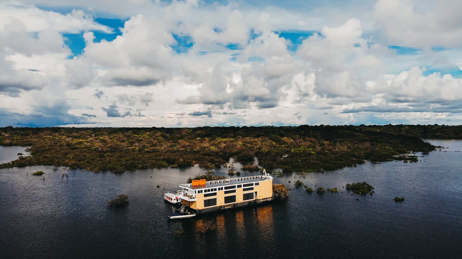 Your floating base camp, deep in the Amazon's drowned forest. Aerial view of the untamed amazon cruise vessel moored among flooded trees and dark water beneath a dramatic cloudy sky