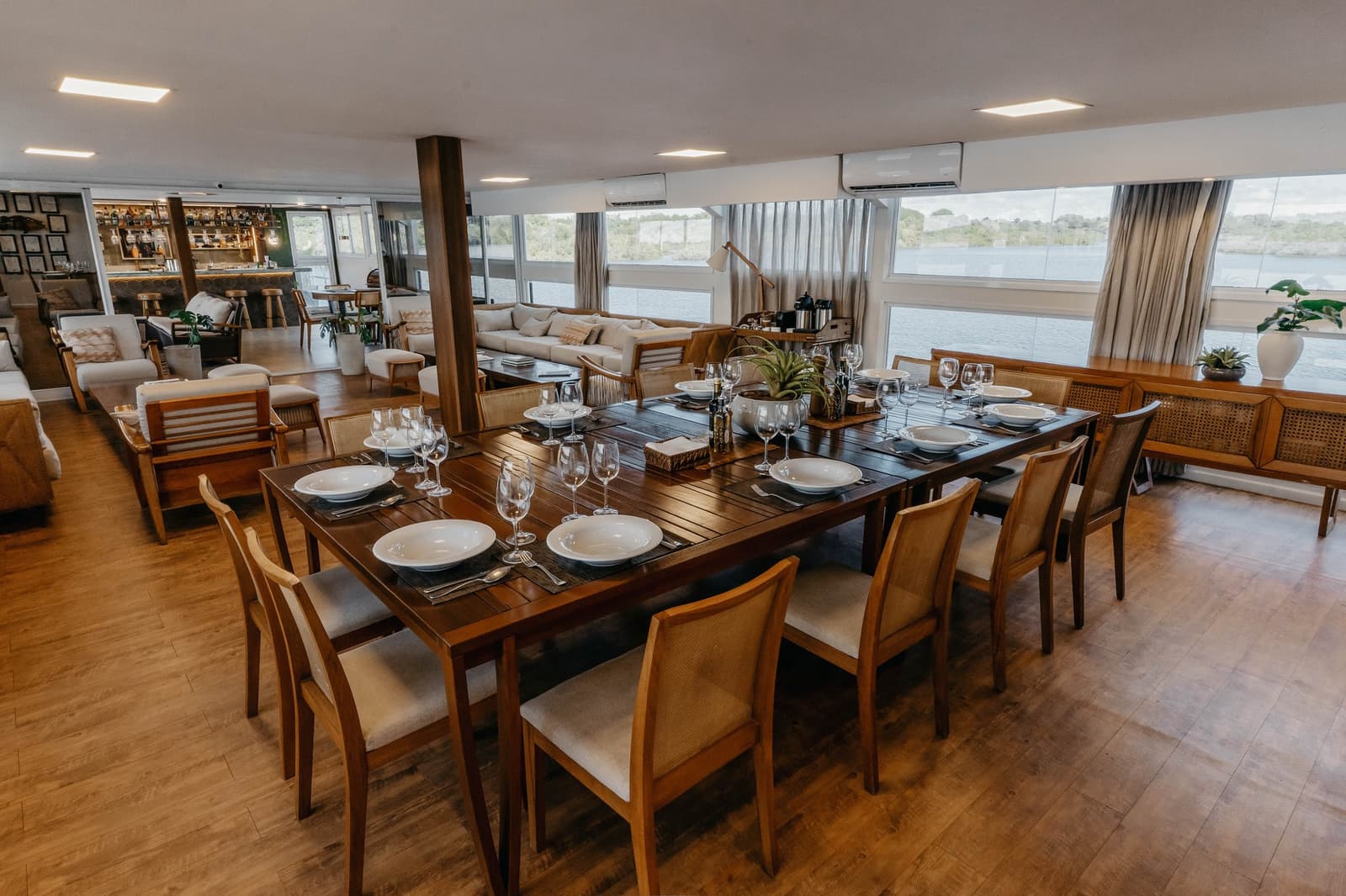 Good meals taste better when the river is right outside the window. Elegantly set dining table with white porcelain, crystal wine glasses and wooden chairs aboard the untamed amazon cruise, with panoramic river views through wide windows and a lounge bar visible in the background