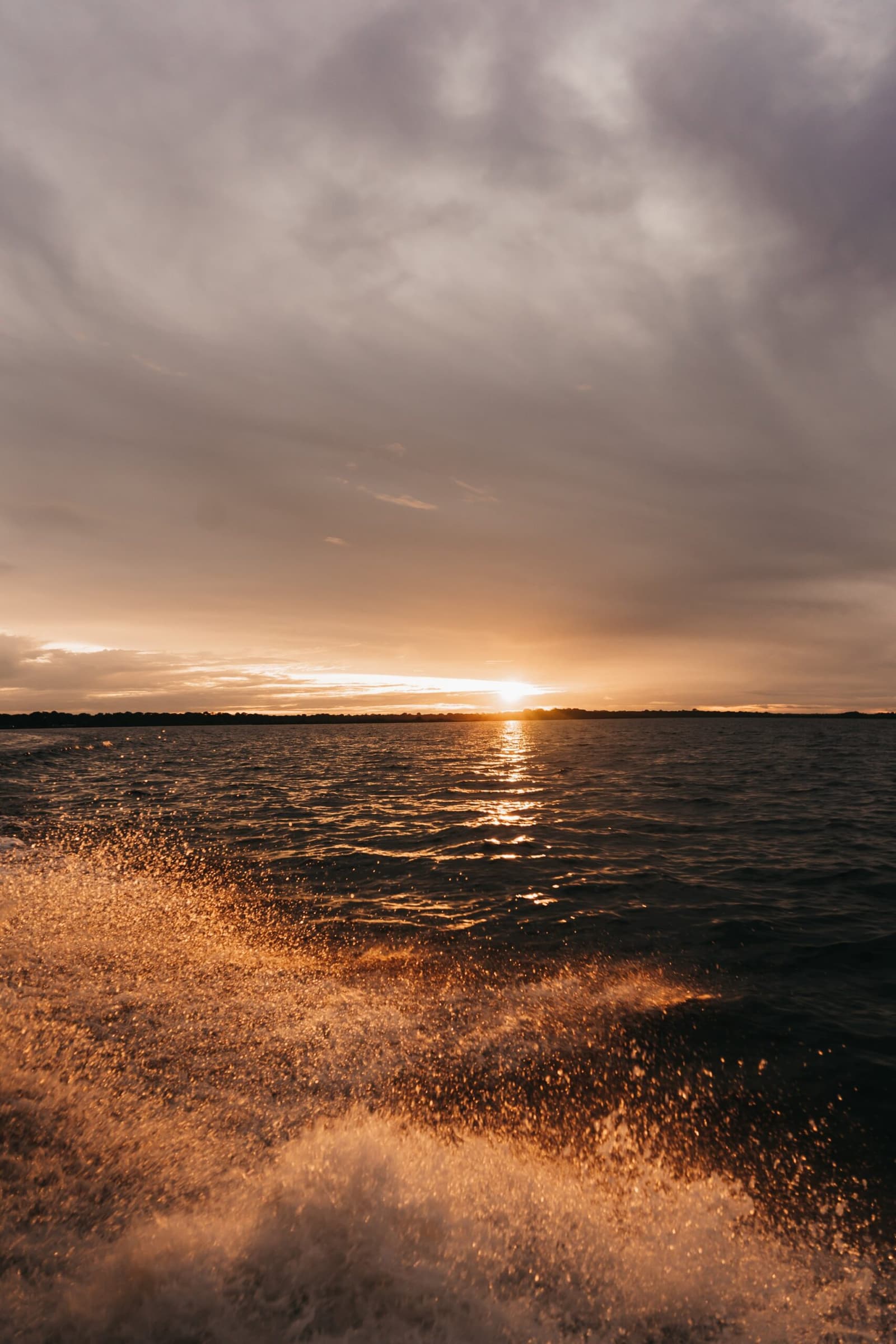 That last hour on the water, when the river turns to liquid copper. Golden boat wake glittering in the foreground as the sun sets low over a wide river on an untamed amazon cruise, with a distant treeline silhouetted beneath a dramatic cloudy sky