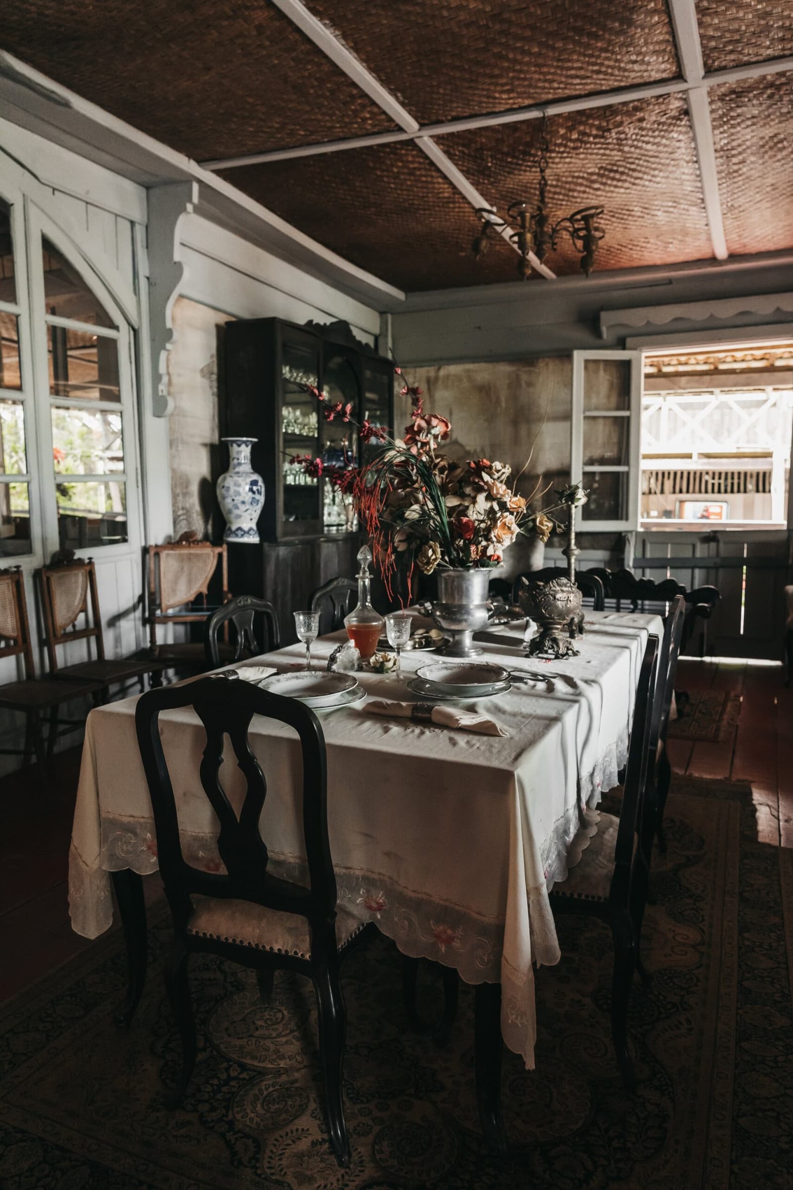 Someone set this table as if guests were expected a century ago. A formally set dining table with white linen, dark carved chairs, antique silverware, crystal glassware and a dried floral centrepiece inside a colonial-era room with a woven ceiling and vintage china cabinet — part of the untamed amazon cruise shore experience