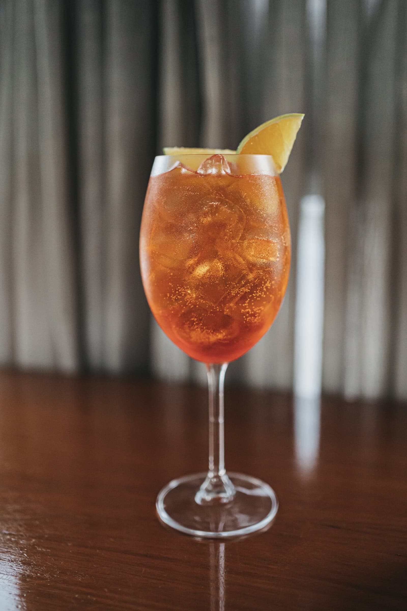 The river can wait five more minutes. An amber iced cocktail in a stemmed wine glass garnished with a citrus wedge, served on a dark wood bar aboard the untamed amazon cruise