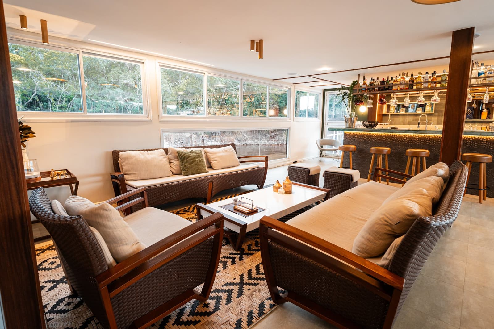 After a day on the river, this lounge earns its keep. The onboard lounge of the untamed amazon cruise featuring woven rattan armchairs, cream cushions, a geometric patterned rug, and a fully stocked bar, with panoramic windows overlooking dense riverside forest