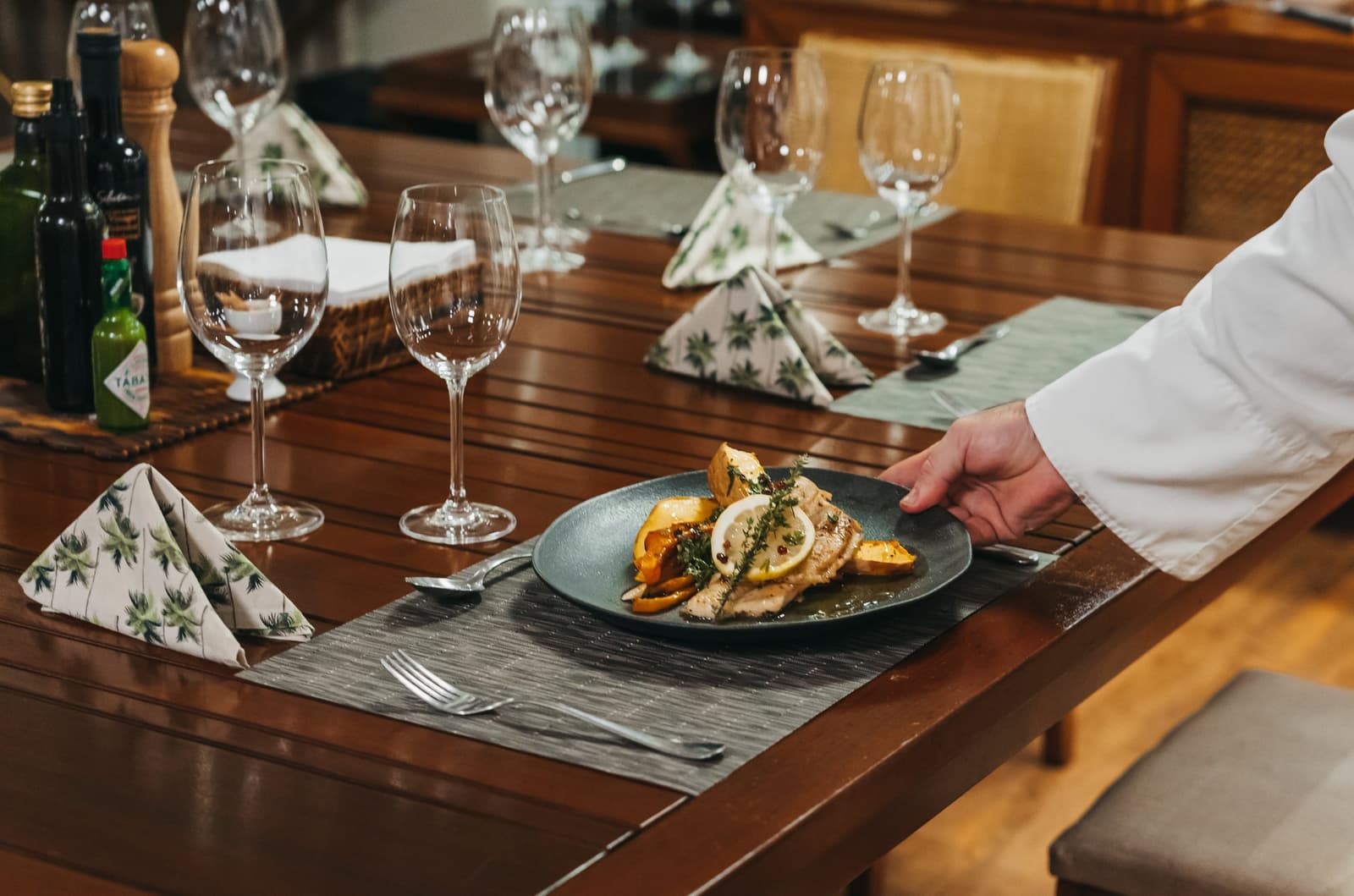 Good food doesn't stop at the riverbank. A chef in a white jacket placing a dark plate of grilled fish with roasted peppers, lemon and fresh herbs on a formally set wooden dining table aboard the untamed amazon cruise
