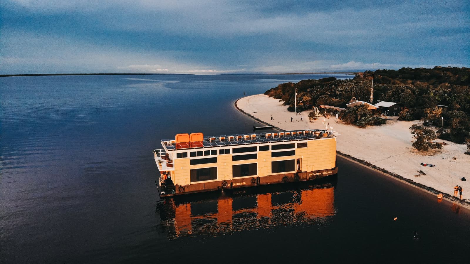 Your floating base camp, right where the river meets the sand. Aerial view of the Untamed Amazon Cruise solar-powered vessel moored beside a white-sand river beach at dusk, with dark water stretching to the horizon and forest-fringed shoreline behind