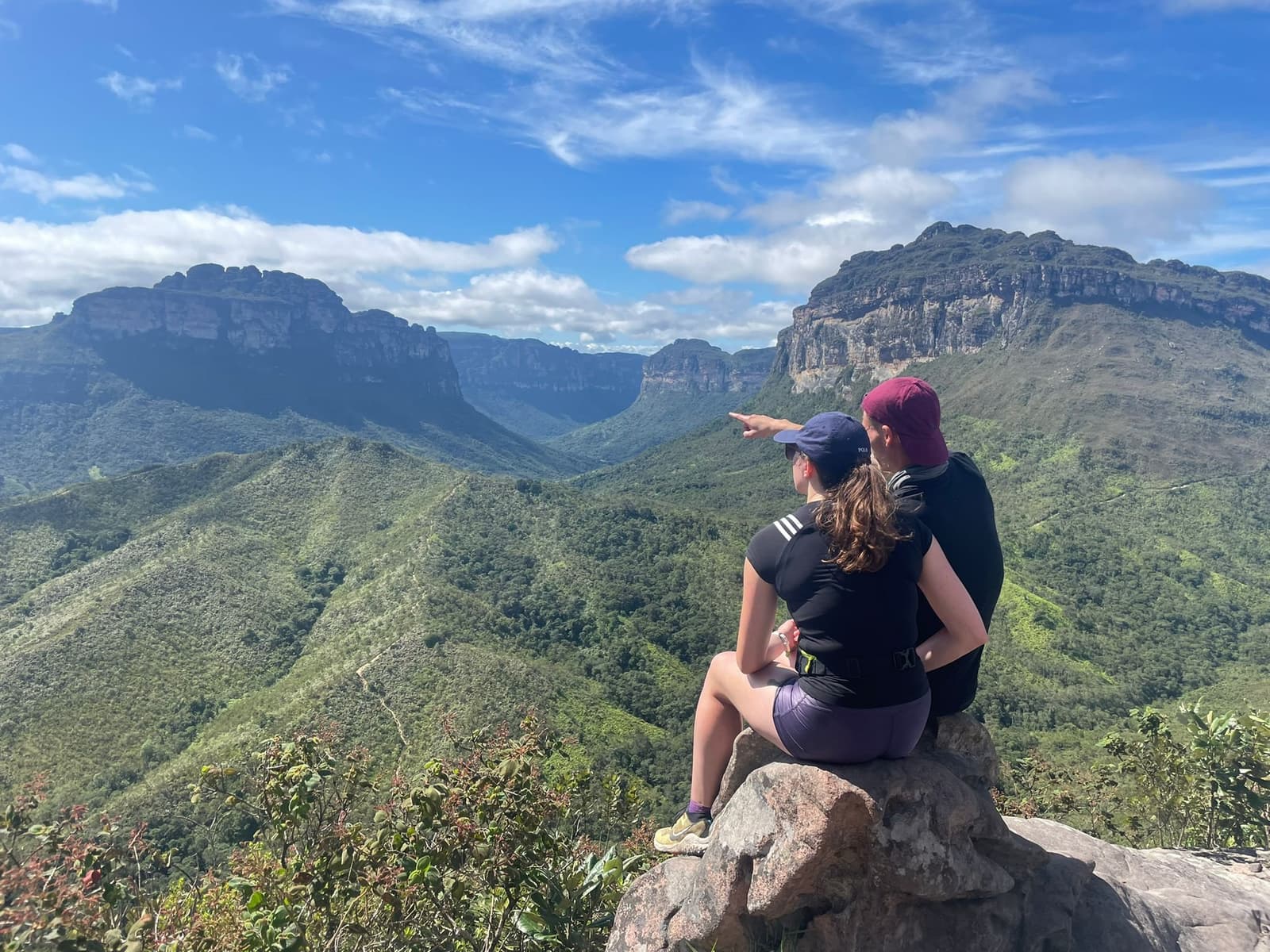 Someone always spots something worth pointing at up here. Two hikers sit on a boulder at a Vale do Pati Trek viewpoint, one pointing toward a sweeping canyon flanked by towering sandstone mesa formations and dense green valley below