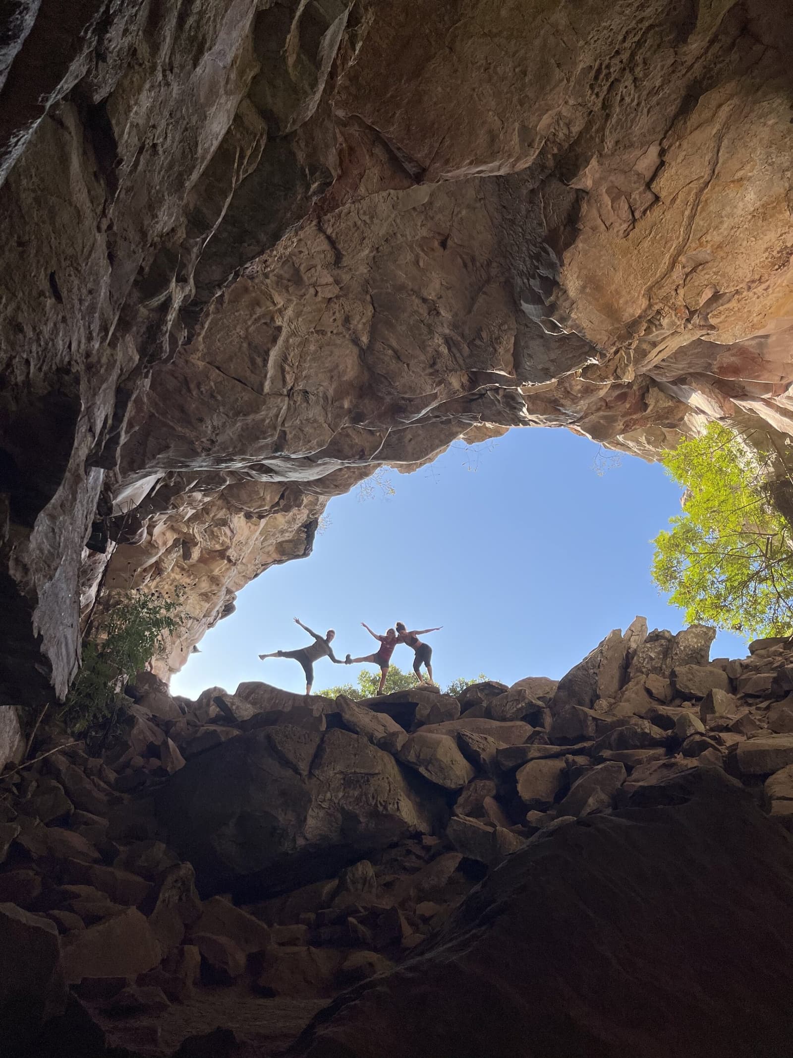 The rock does its part; they do the rest. Two trekkers striking balance poses on boulders framed by a massive sandstone cave arch on the Vale do Pati Trek, blue sky and green treetops visible beyond the opening