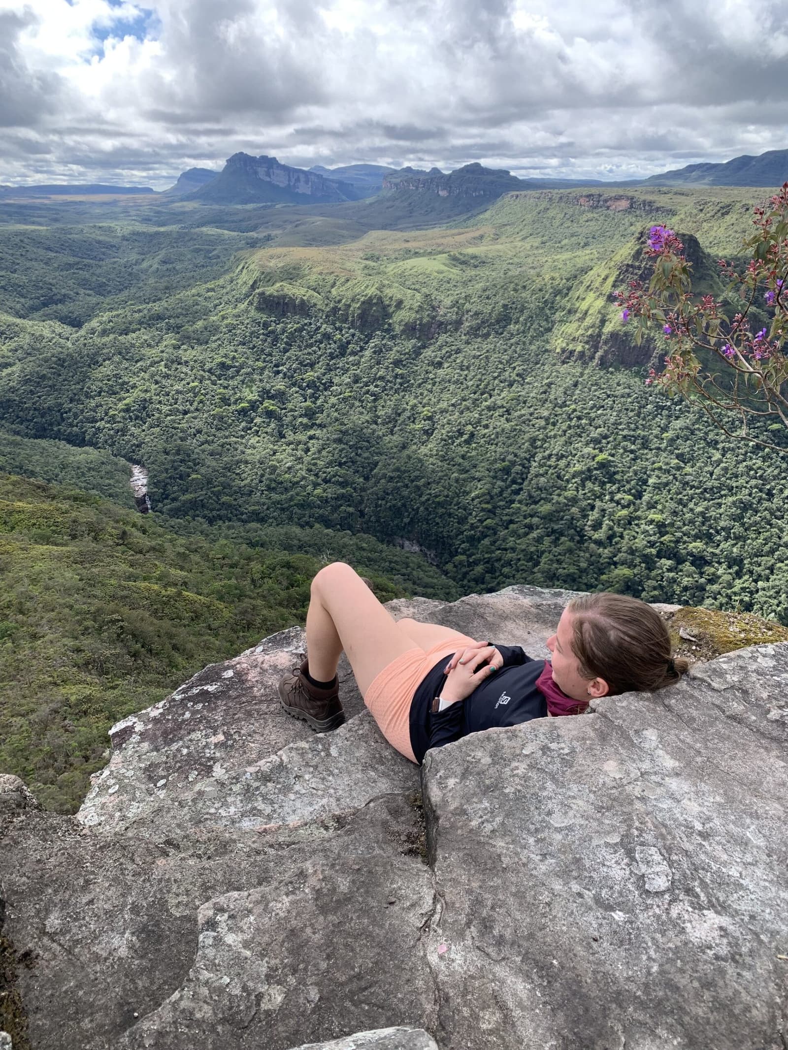 Best office view in Bahia, and the chair is a boulder. A hiker lies on a rocky cliff edge on the Vale do Pati Trek, gazing over a vast forested canyon with flat-topped sandstone plateaus and a distant waterfall under a cloudy sky in Chapada Diamantina