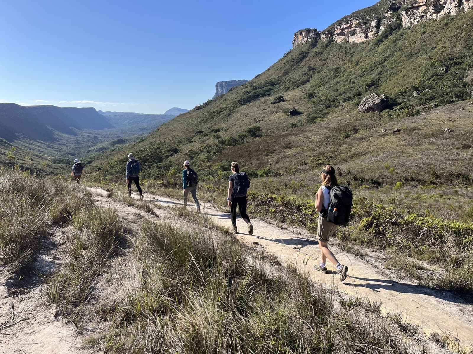 The valley opens up slowly, and that first long view earns every step. Five backpackers hiking single-file along a sandy stone trail on the Vale do Pati Trek, with sweeping valley views and towering sandstone escarpments under a clear blue sky