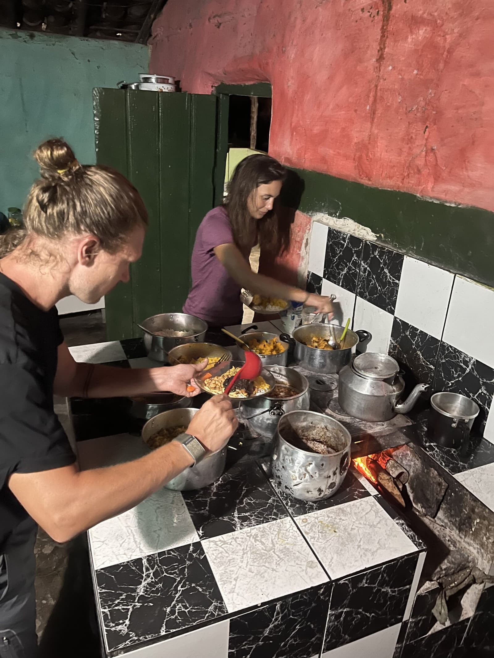 Dinner earns its place after a long day on the trail. Two trekkers serving themselves from multiple pots simmering over a wood-fired stove in a rustic tiled kitchen during the Vale do Pati Trek homestay dinner