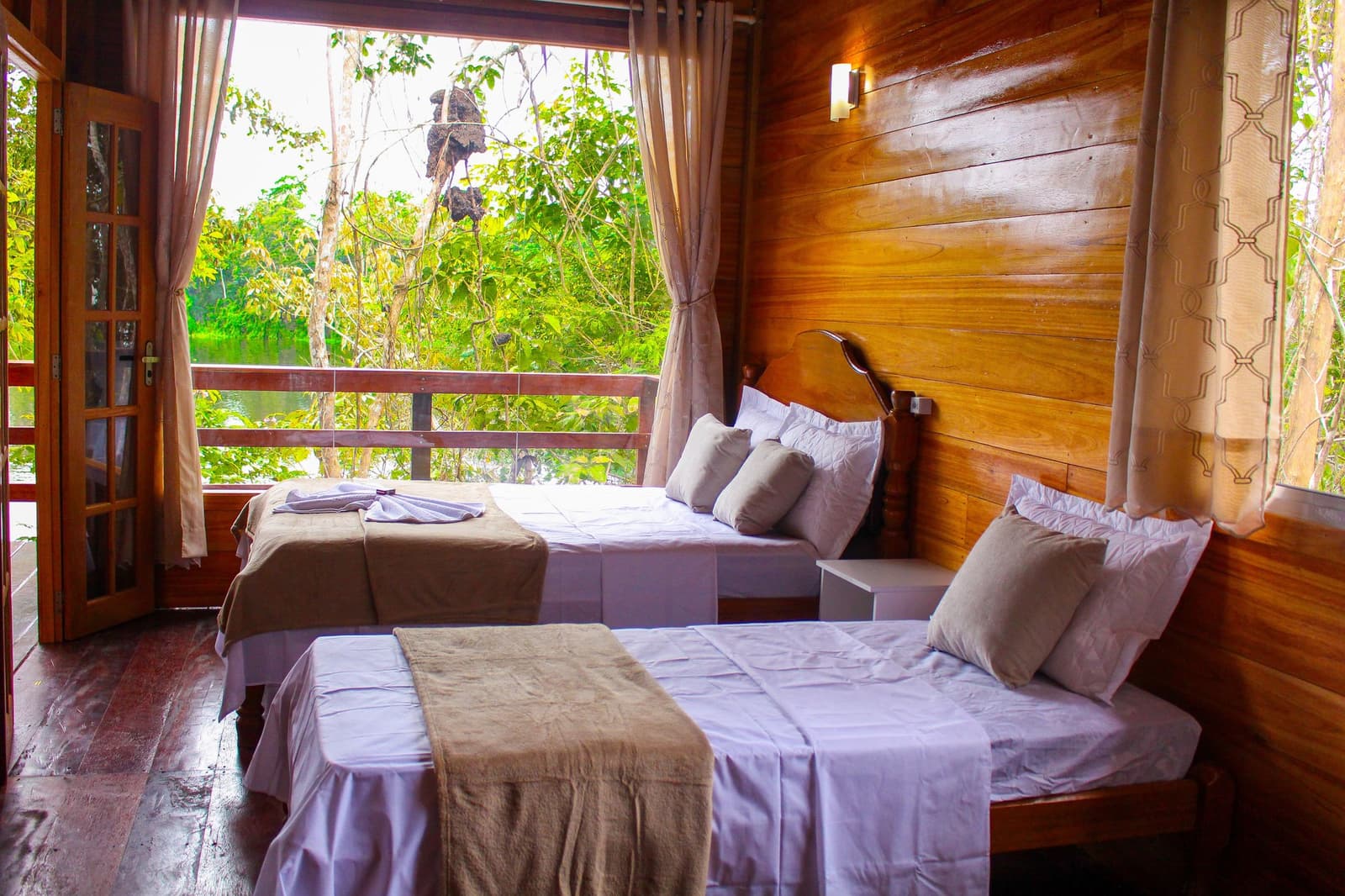 Sleep with the forest on your doorstep, the river just a little closer. Twin-bed room in an amazon jungle lodge with warm timber walls, white linen, and an open balcony door framing dense green forest and a calm river beyond