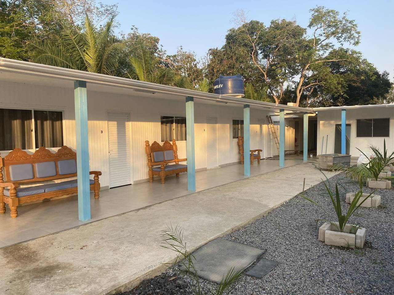 Carved benches on the veranda, trees right behind you. Comfort earned. A row of numbered guestroom doors along a covered veranda at an amazon and pantanal tour lodge, with carved wooden benches outside each room and tall trees visible behind the white-painted building