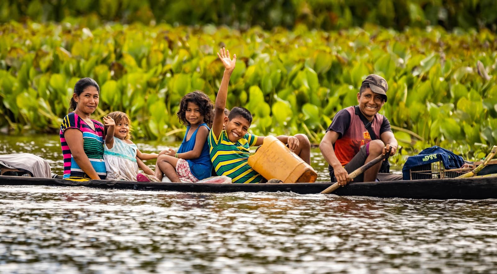 The warmest welcome on the river, no reservation needed. Photo: GummyBone A smiling local family of five paddling a wooden canoe on a dark river during an amazon and pantanal tour, with a boy waving and lush aquatic vegetation in the background