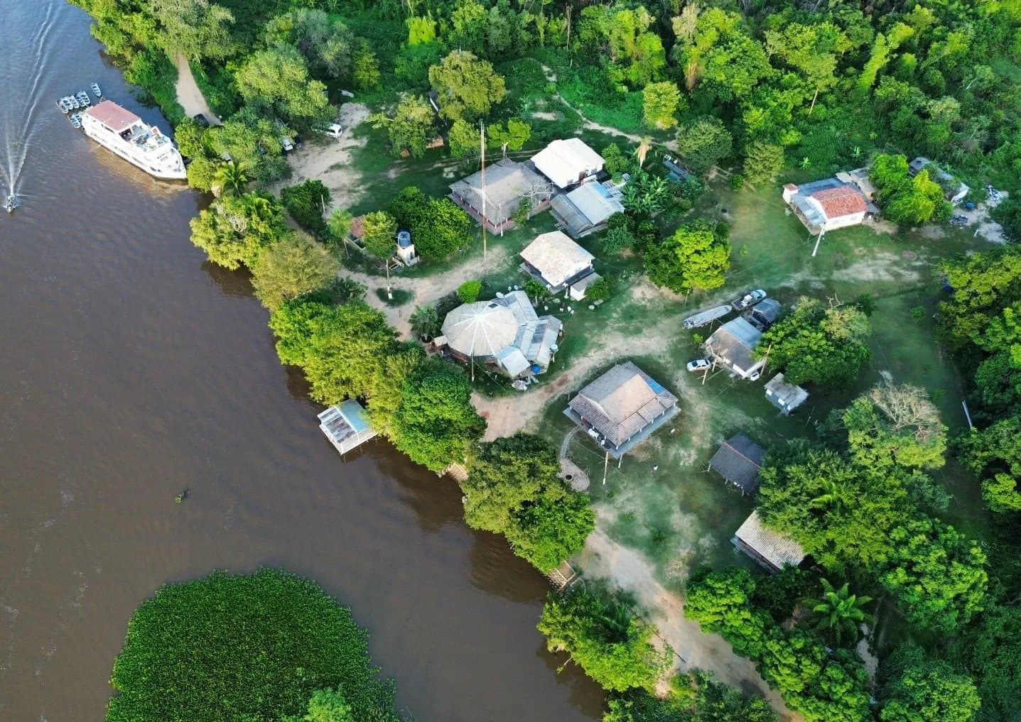 Tucked into the trees, closer to the river than it looks on the map. Aerial view of a small riverside settlement on an amazon and pantanal tour, with thatched and tiled-roof buildings nestled among dense green trees along a brown river, a multi-deck boat moored at the bank