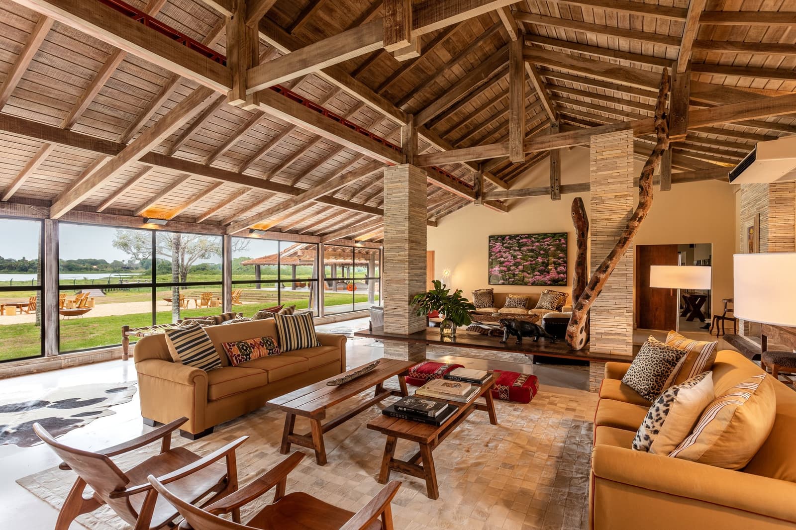 The kind of common room that makes you forget there are jaguars waiting outside. Open-plan lodge lounge at Caiman on a luxury Pantanal and Amazon trip, featuring exposed hardwood beam ceilings, amber sofas with animal-print cushions, a driftwood sculpture, and floor-to-ceiling windows overlooking green wetland floodplains
