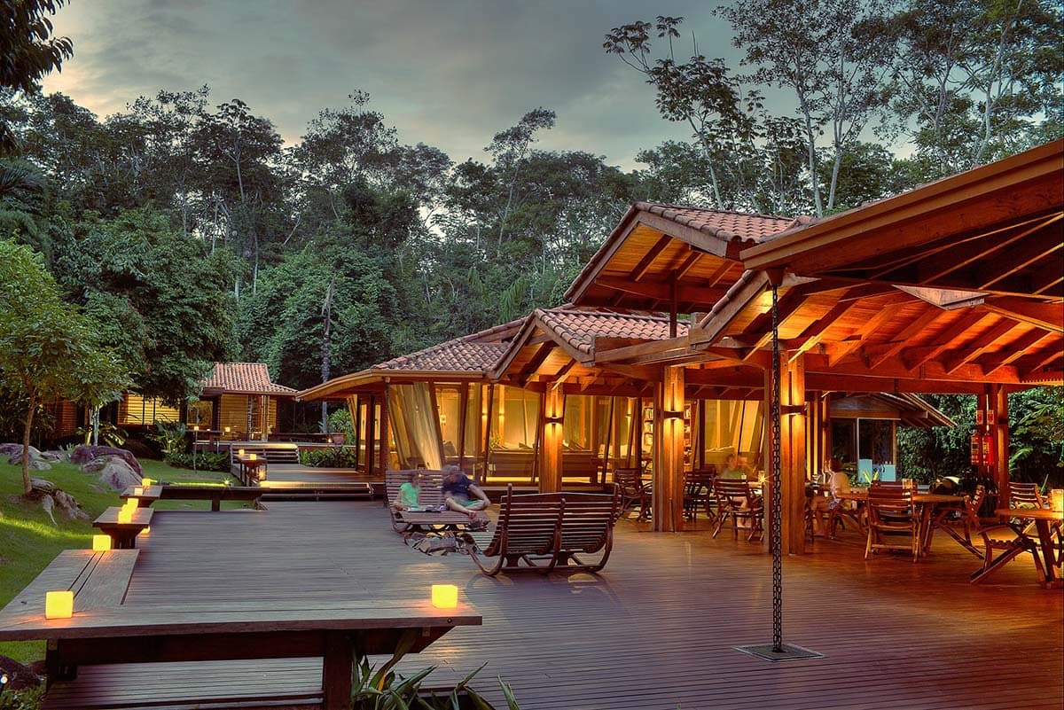 Candles out, forest close, and someone already in a rocking chair. That is a good sign. Photo: Samuel Melim Warm-lit wooden deck and open pavilions of a luxury pantanal and amazon trip lodge at dusk, with candles glowing on low platforms, guests relaxing in rocking chairs, and dense rainforest rising behind the buildings