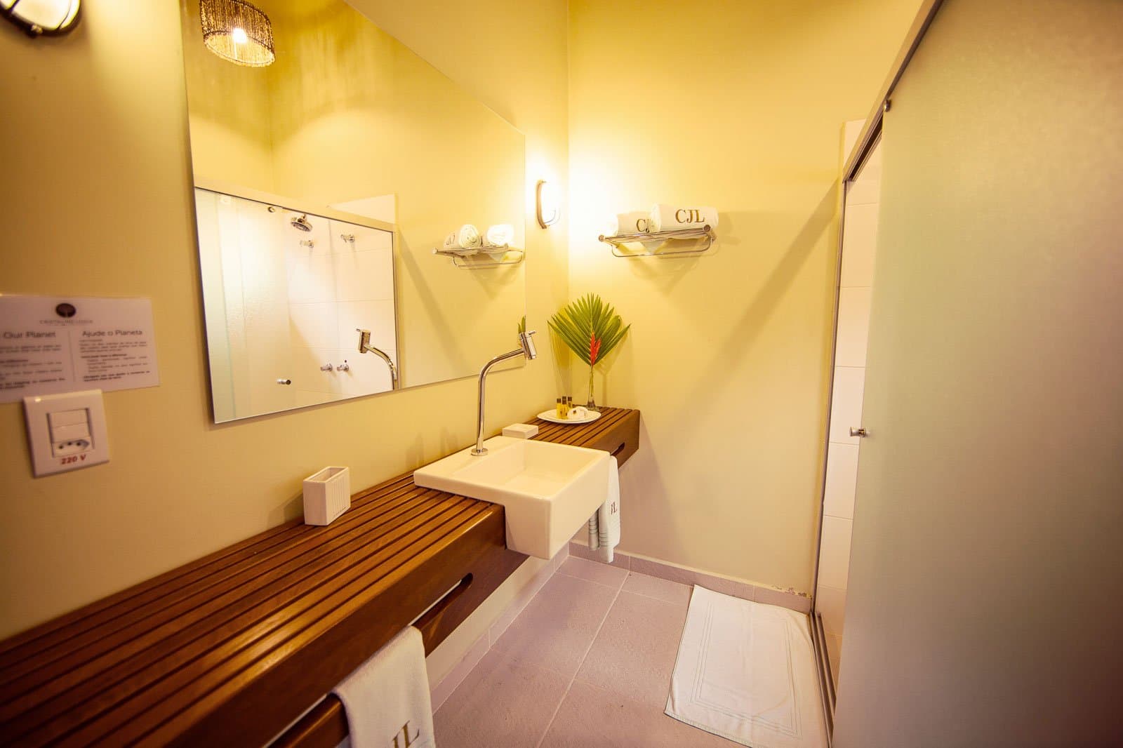 Teak slats, monogrammed towels, a heliconia bloom. The wild can wait five more minutes. Photo: | SamuelMelim Photography Lodge bathroom on a luxury Pantanal and Amazon trip featuring a square vessel sink on a teak slatted vanity, warm yellow walls, monogrammed towels, and a tropical heliconia stem in a vase
