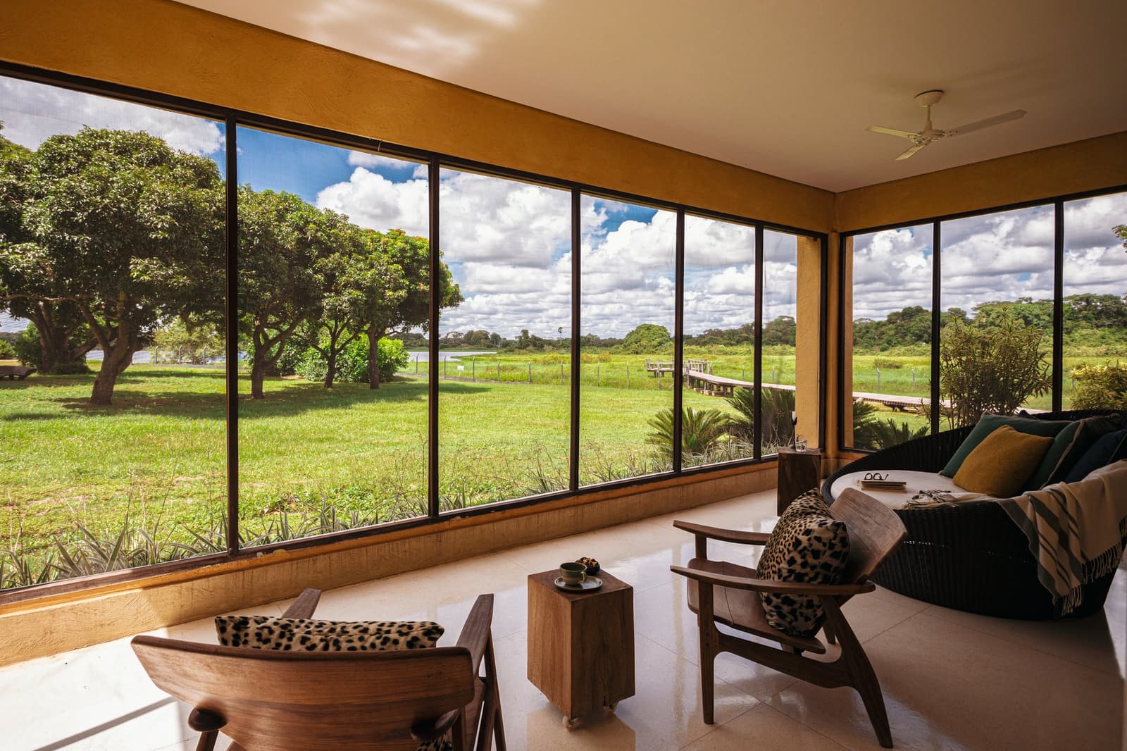 The best seat in the Pantanal comes with a ceiling fan and a coffee. Photo: Felipe Castellari Screened lounge area of a luxury Pantanal and Amazon trip lodge, with wooden armchairs and animal-print cushions facing floor-to-ceiling windows overlooking a green floodplain, wooden boardwalk, and partly cloudy sky