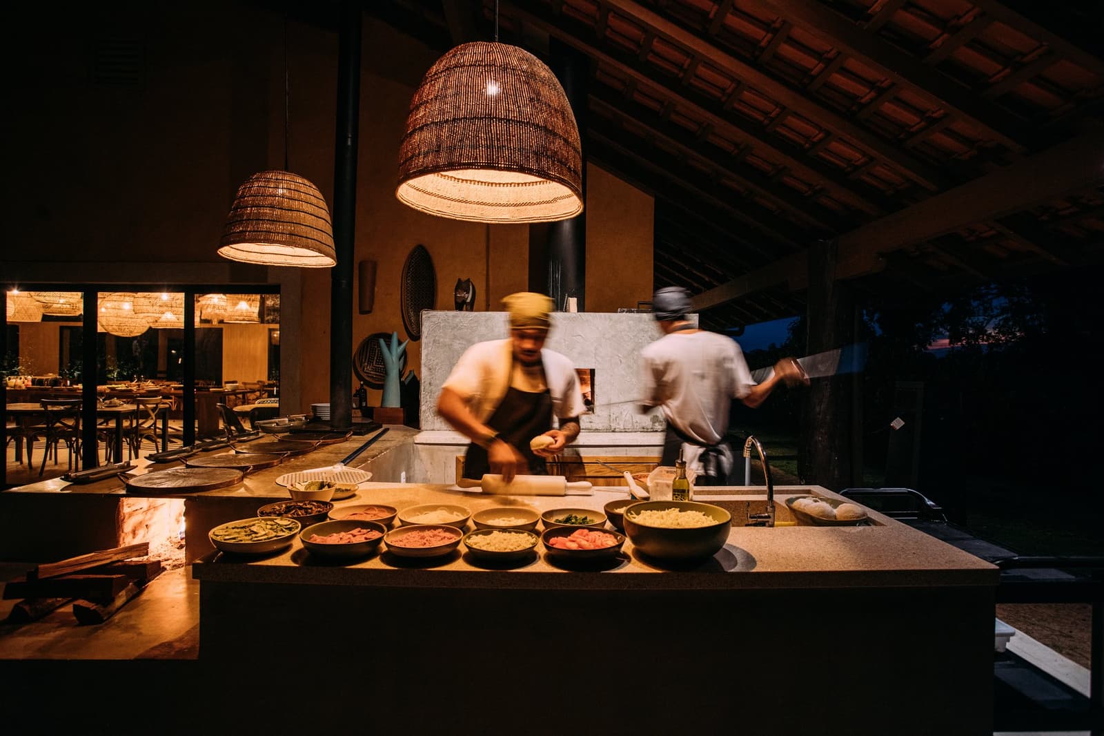 Dinner doesn't happen to you here, it's made right in front of you, fast hands and all. Two chefs in motion at an open-air wood-fired kitchen station on a luxury Pantanal and Amazon trip, bowls of fresh ingredients lined along a stone counter under warm wicker pendant lights at dusk