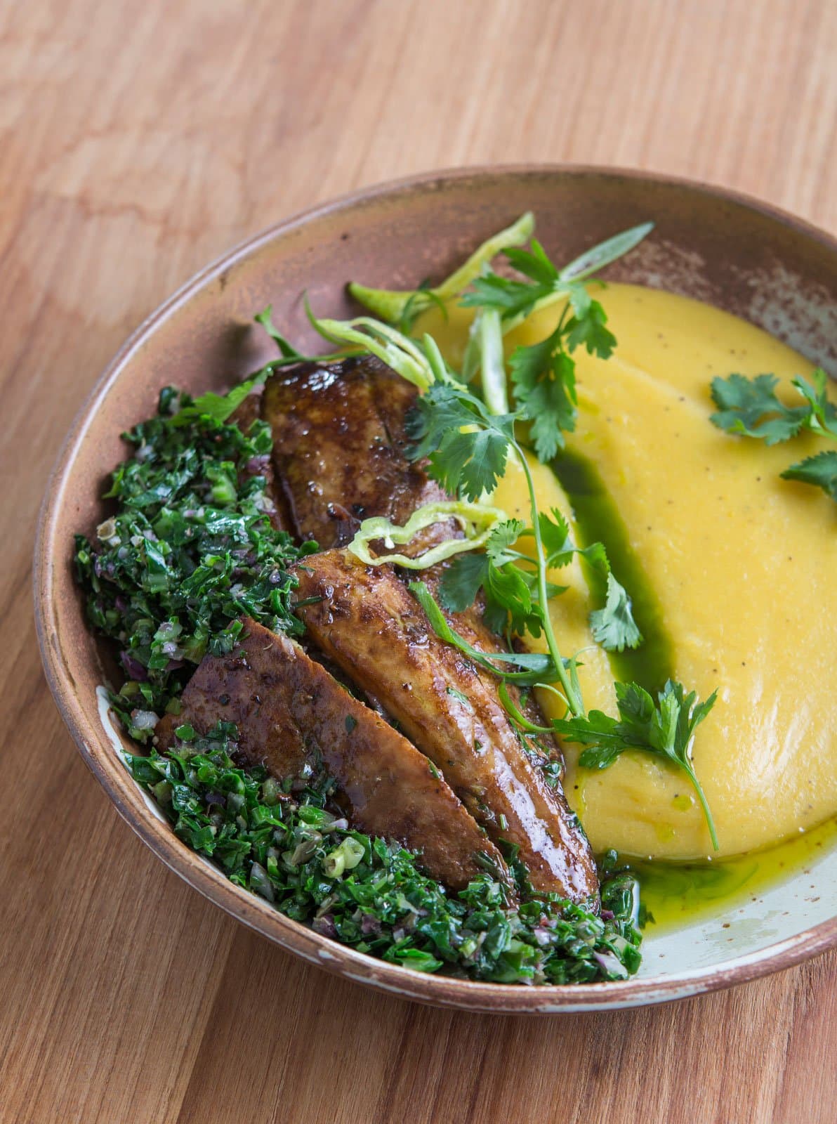 Lodge cooking that treats local ingredients like the headliners they are. Photo: Rjcastilho A rustic ceramic bowl served on a wooden table, featuring caramelized pan-seared fish fillets over a chopped herb salsa, alongside a smooth golden squash or corn purée garnished with fresh cilantro and sliced spring onion, part of the lodge dining experience on a luxury Pantanal and Amazon trip