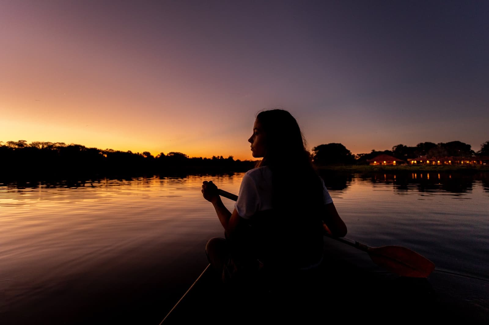 Sunset moves fast out here. Paddle in hand, nowhere else to be. Silhouette of a woman holding a paddle in a canoe on a calm river at sunset during a luxury Pantanal and Amazon trip, with a warm orange and purple sky reflecting on the still water and lit lodge buildings visible on the far bank
