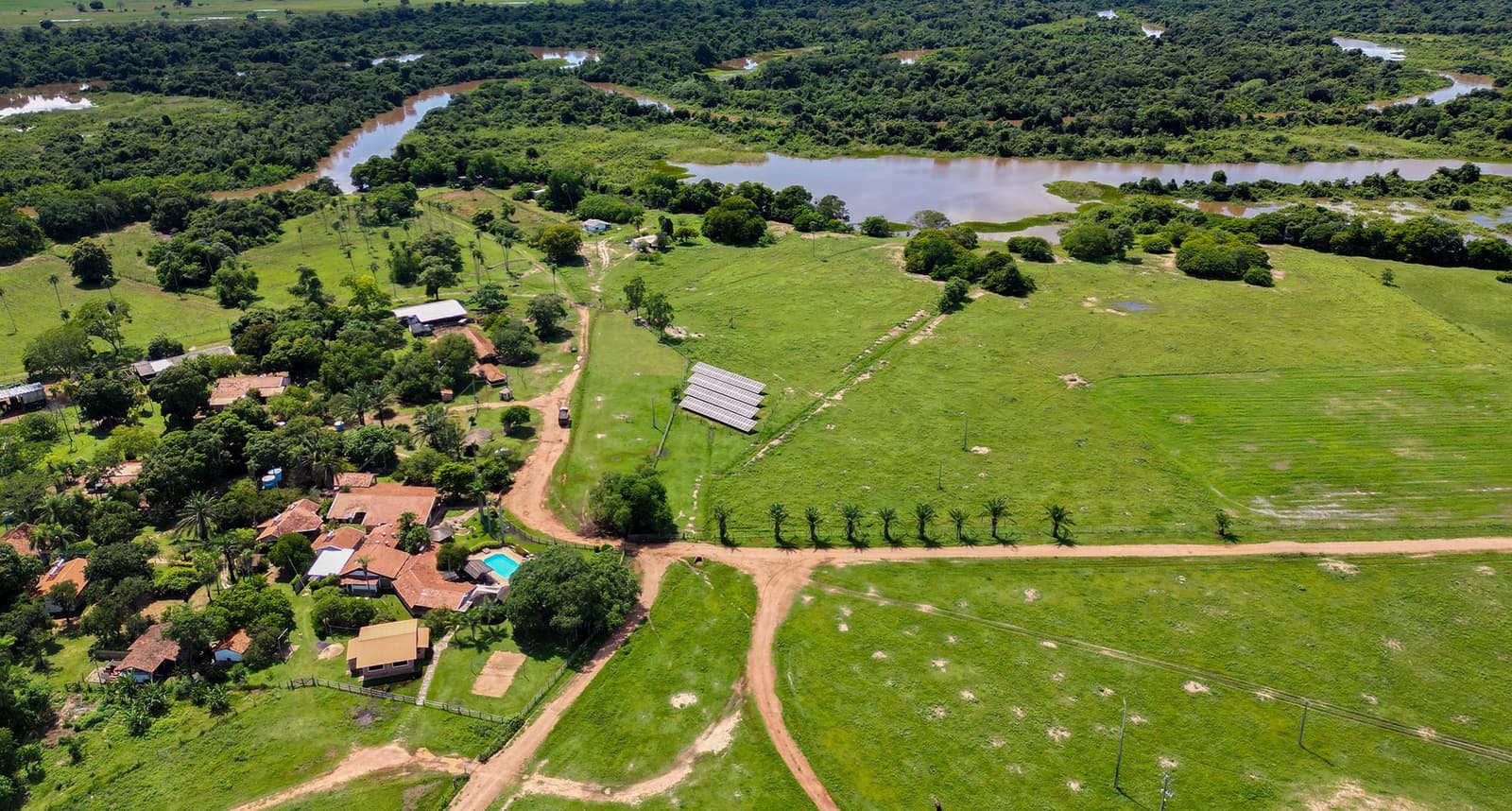 Solar panels next to the cowpaths. Eco-lodges in the Pantanal have come a long way. Aerial view of a Pantanal lodge with solar panels, red-tiled buildings, a pool, and dirt tracks cutting through open green fields beside a winding river and gallery forest on a pantanal bonito tour