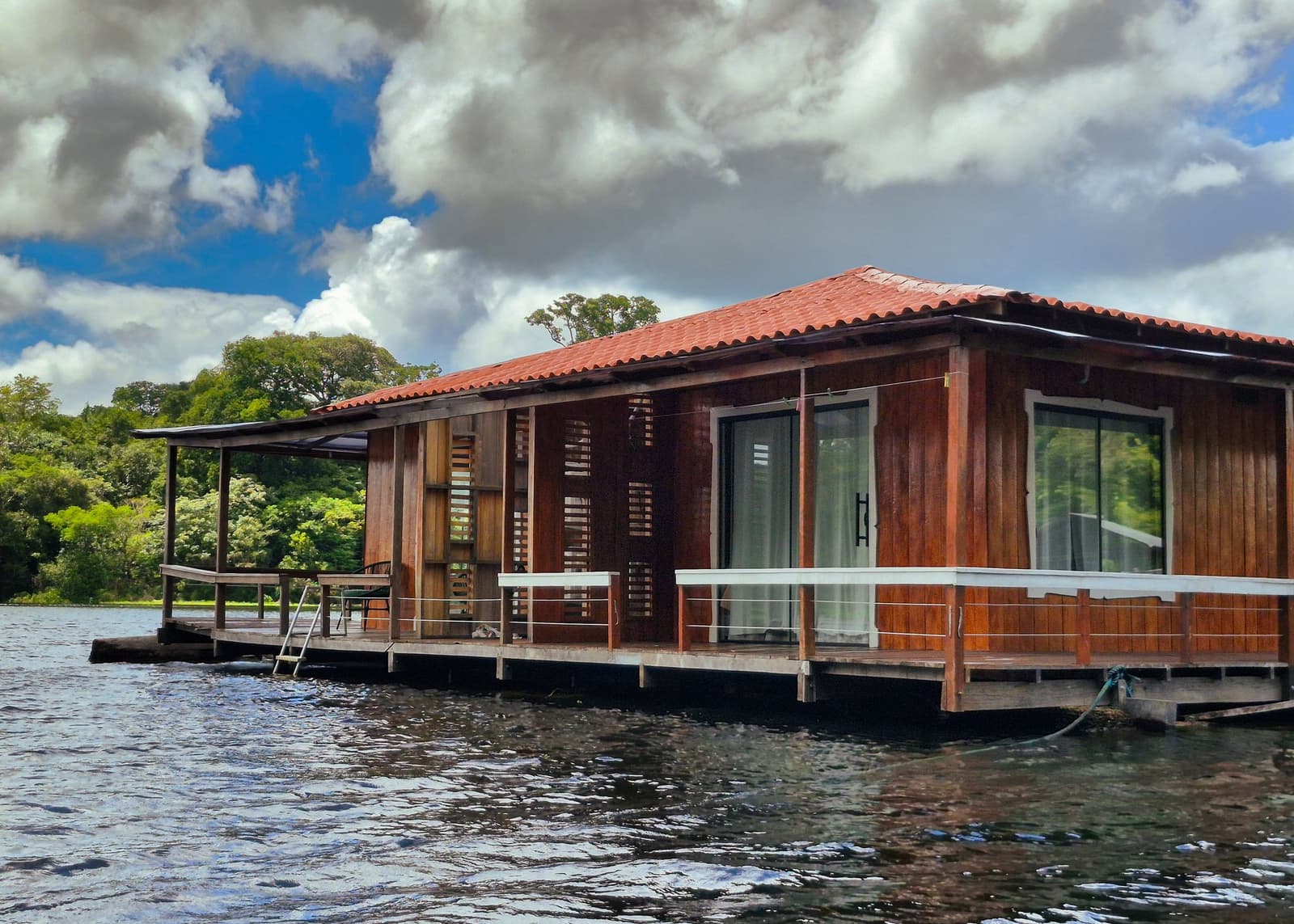 Your room on the water, the forest just a rope's length away. A wooden floating lodge with a red-tiled roof and wraparound deck sits on dark river water, surrounded by dense green forest under a partly cloudy sky — accommodation on an amazon and pantanal tour
