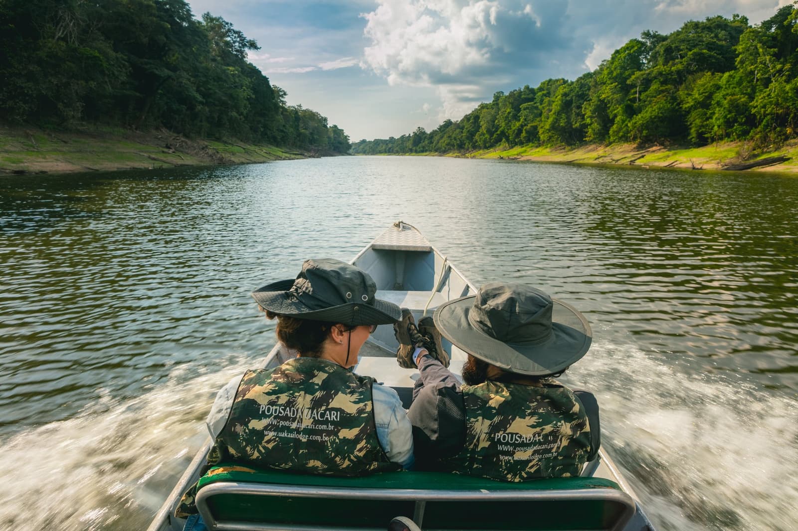 The river doesn't show you everything at once. That's the whole point. Photo: Guilherme Gomes Two guests in camouflage life vests and wide-brim hats sit at the bow of a speedboat on an amazon rainforest ecolodge trip in brazil, pointing ahead as the boat cuts through a calm dark-water river flanked by dense jungle on both sides