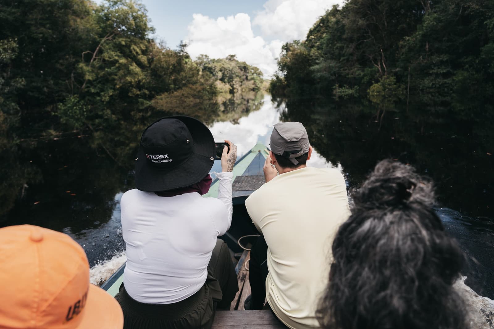 That channel ahead looks narrow. It opens up, it always does. Four travellers seated aboard a wooden boat on a luxury Amazon cruise in Brazil, navigating a narrow blackwater channel flanked by dense rainforest, one passenger photographing ahead with a smartphone