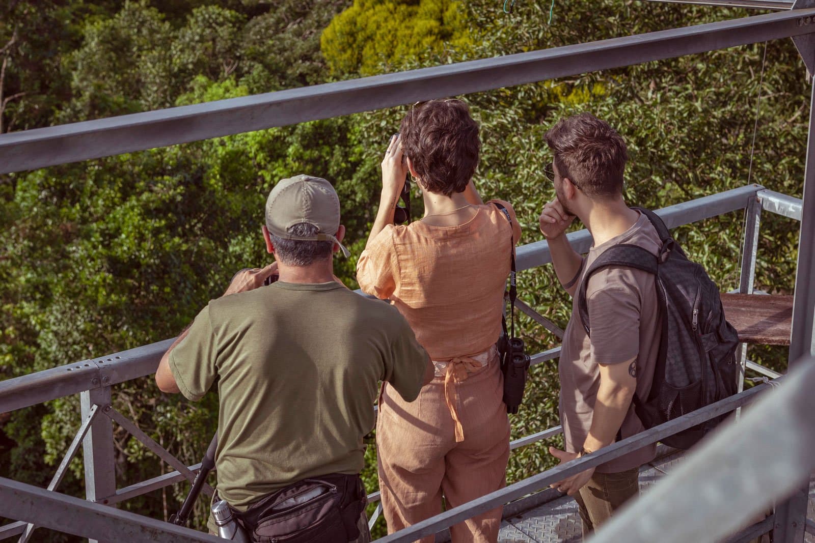 Binoculars up, arguments over the bird ID coming shortly. Photo: Samuel Melim Three visitors on a metal canopy tower scan the forest canopy with binoculars on a luxury Pantanal and Amazon trip, surrounded by dense green treetops in bright sunlight