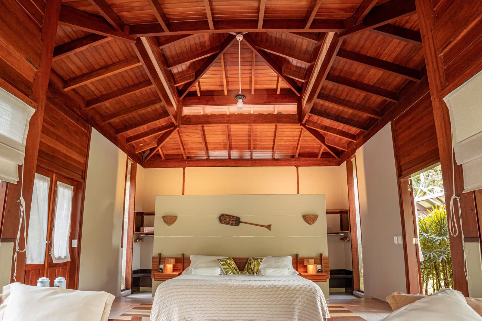 That ceiling does something to you the moment you walk in. Photo: Samuel Melim Spacious lodge bedroom on a luxury Pantanal and Amazon trip, featuring a soaring timber-framed ceiling, white bedding with leaf-print cushions, warm candlelight on wooden nightstands, and a decorative paddle mounted above the headboard