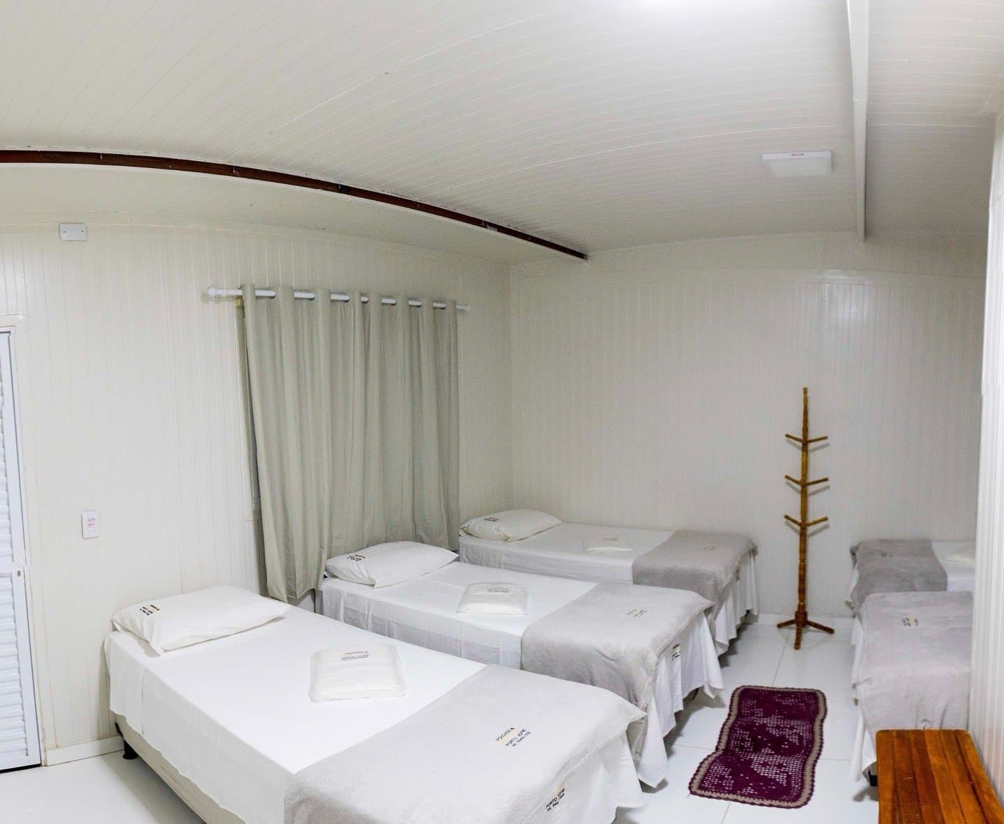 Simple, tidy, and ready for people who came to sleep well before an early departure. Clean shared bedroom with four single beds dressed in white linen, a wooden coat stand, and a curtained window inside an amazon and pantanal tour lodge