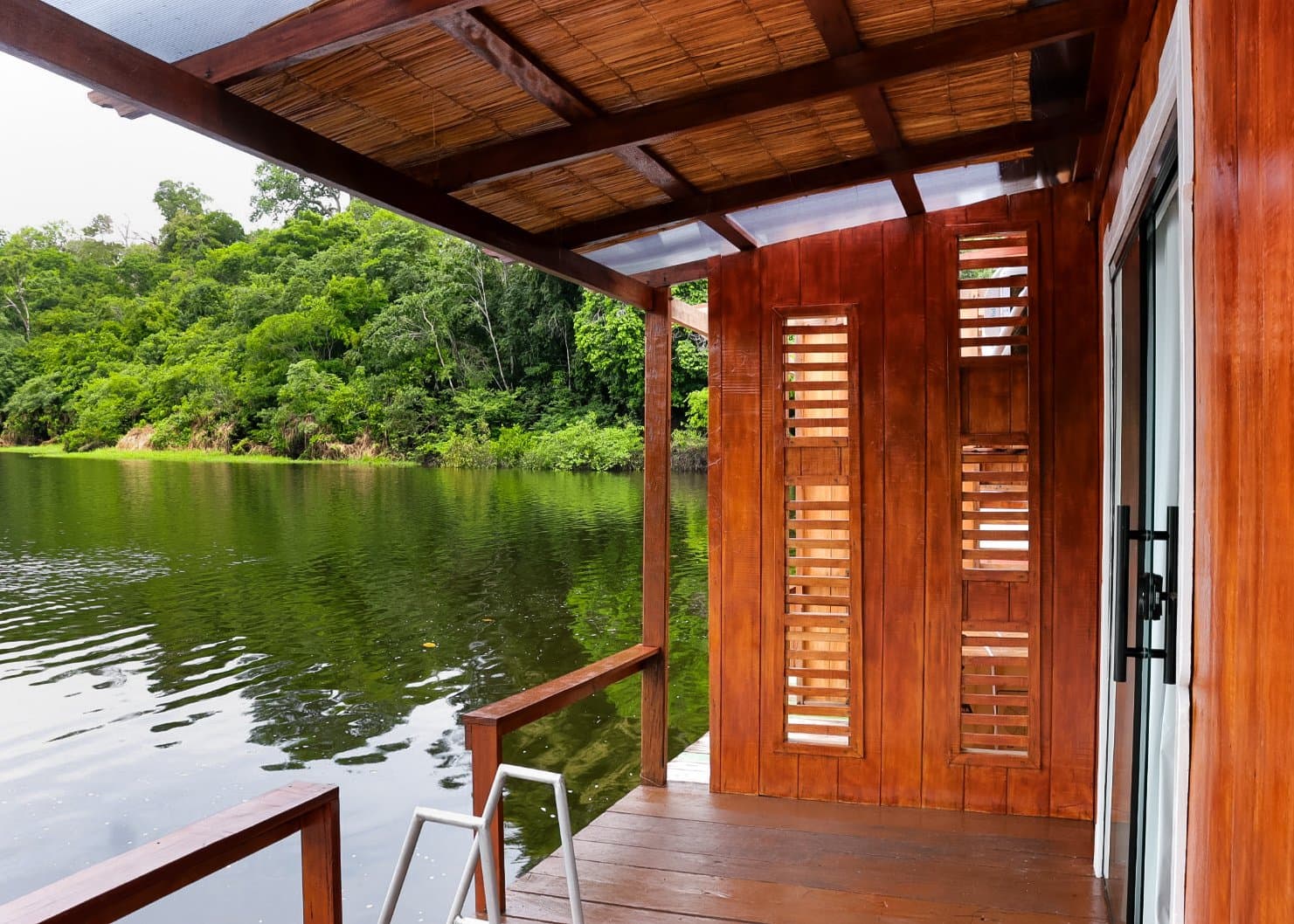 The forest is literally across the water. Sit down, it can wait. Covered wooden deck of an amazon jungle lodge overhanging a dark, still river, with louvred hardwood shutters and a thatch-and-timber ceiling framing dense green rainforest on the opposite bank