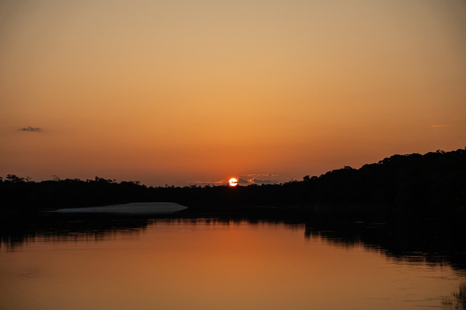 Some sunsets just stop the conversation cold. A deep orange sunset over a calm Amazon river, the sun touching the treeline silhouette and reflected in the still water below, with a white sandbar visible on the left bank — seen from a luxury Amazon cruise in Brazil