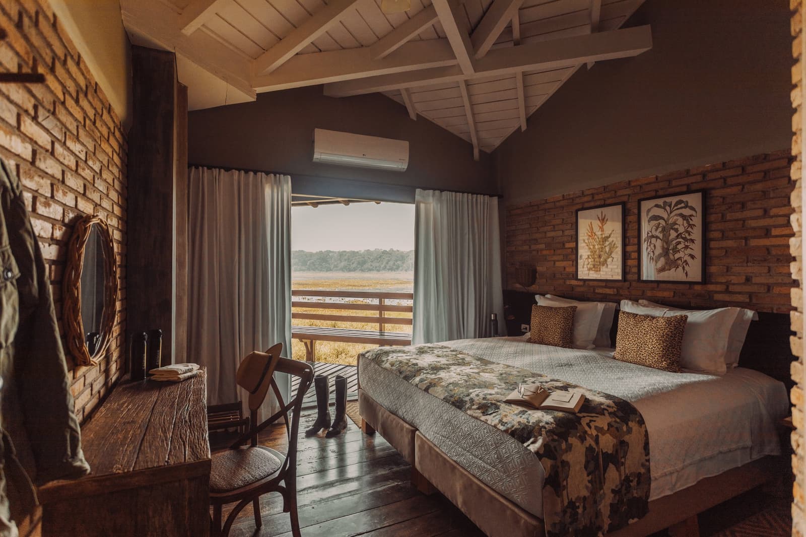 Rubber boots at the ready, book on the bed. Safari life suits itself. Photo: Felipe Castellari Rustic-luxe double room on a luxury Pantanal and Amazon trip, with exposed brick walls, dark wood floors, leopard-print pillows, rubber boots by the balcony door, and a wide window framing open wetland and treeline beyond