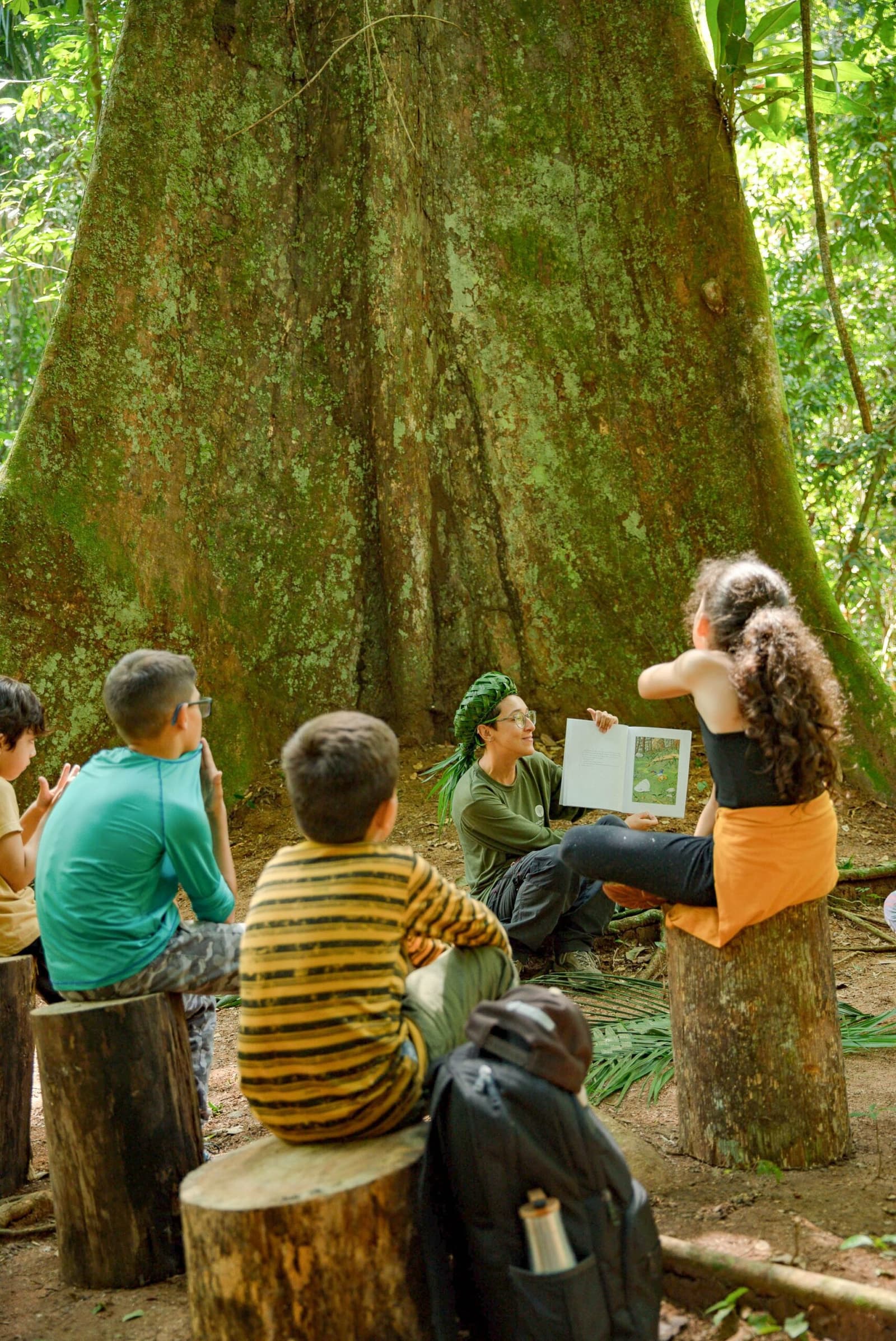 The biggest classroom has no walls, no desks, and one very good tree. A guide sits smiling against the massive moss-covered buttress roots of a large rainforest tree, reading from an illustrated book to a small group of children seated on log stumps during a luxury Pantanal and Amazon trip nature activity