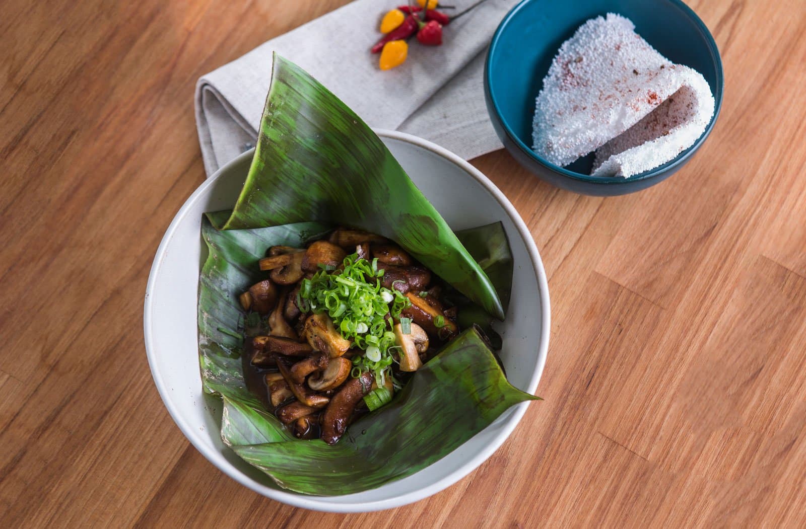 Forest ingredients, plated with a little ceremony and a lot of intention. Photo: Rjcastilho Sautéed mushrooms topped with sliced spring onions served in a banana leaf inside a white bowl, alongside a blue bowl of farofa dusted with chili, fresh peppers on a linen napkin in the background, part of a luxury Pantanal and Amazon trip dining experience