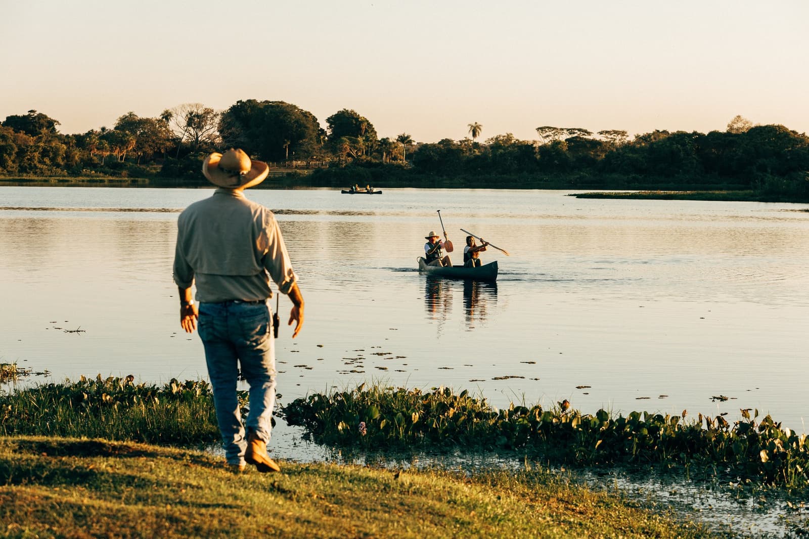 The guide on shore, the guests on the water. Someone has to keep watch. A guide in a wide-brimmed hat watches from the grassy shore as two guests paddle a canoe across a calm Pantanal lagoon at golden hour, with tree-lined banks reflected in the still water during a luxury Pantanal and Amazon trip