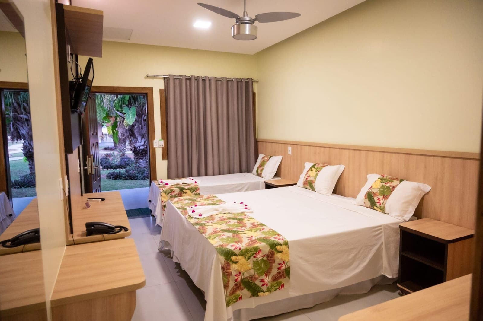 Tropical prints, a ceiling fan, and a garden just outside the door. Comfort sorted. Triple room at a Pantanal ranch-lodge on an amazon and pantanal tour, with two double beds dressed in tropical-print runners, ceiling fan, wall-mounted TV, and an open door revealing a lush garden