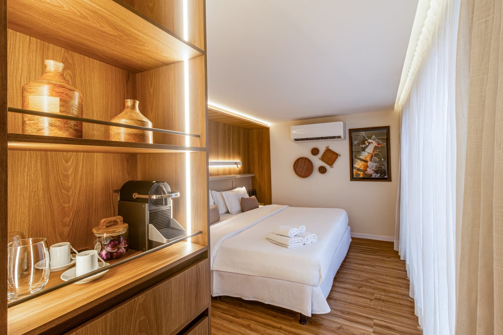 Coffee ready before you even think about asking for it. Photo: STUDIO CREATE A well-appointed cabin on a luxury Amazon cruise in Brazil, featuring a double bed with fresh white linens and folded towels, a backlit wooden shelving unit with a capsule coffee machine, ceramic cups, and decorative clay vases, woven basket wall art, and sheer curtains beside a wood-panel headboard