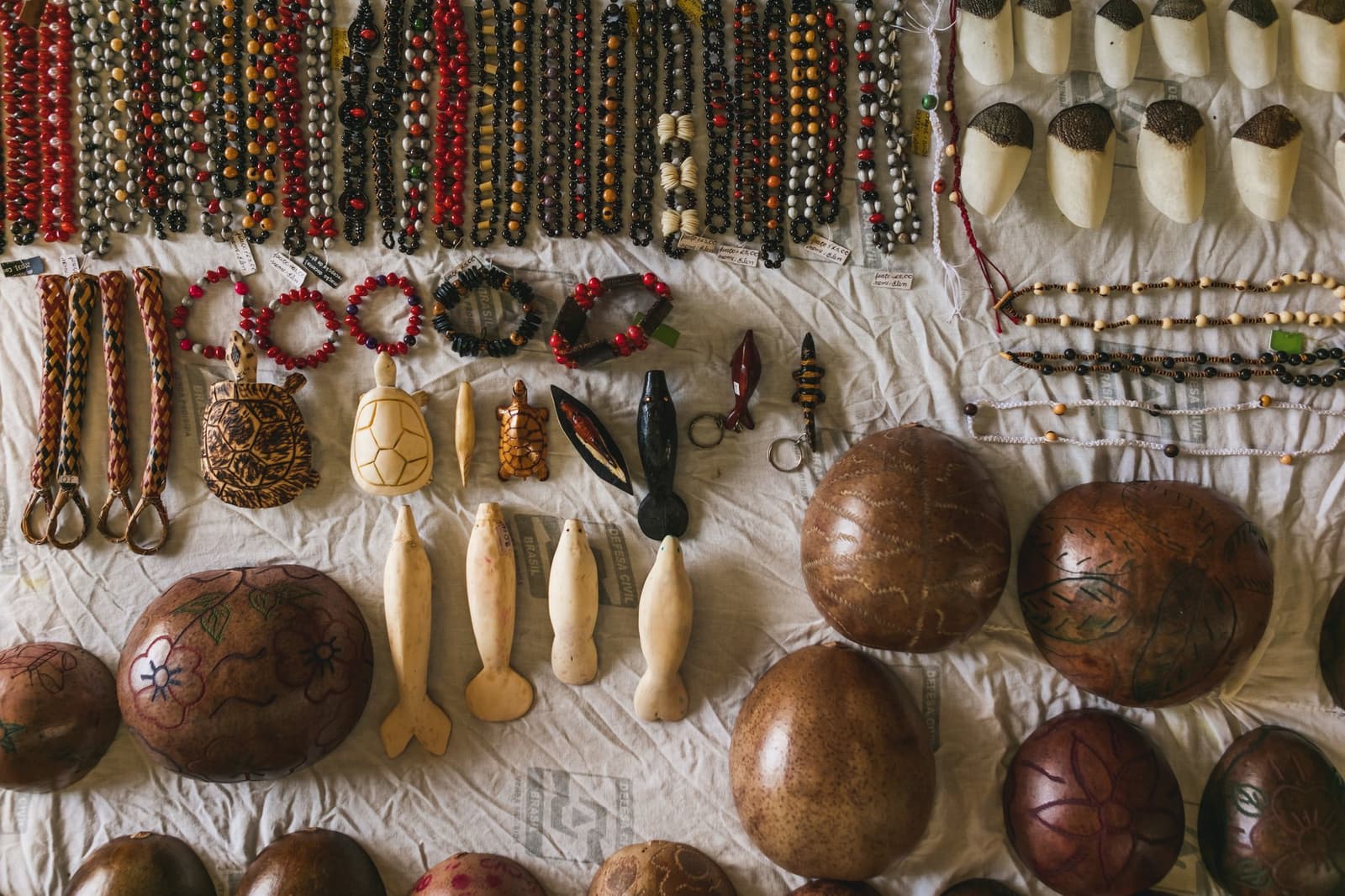 Made by hand, priced in pencil on paper, take your time choosing. Photo: Guilherme Gomes Flat-lay of Amazonian handmade crafts on a white cloth during an amazon rainforest ecolodge trip in brazil, including seed-bead necklaces and bracelets in red, black and orange, carved turtle figurines, dolphin-shaped ornaments, woven keychains, and painted gourd bowls