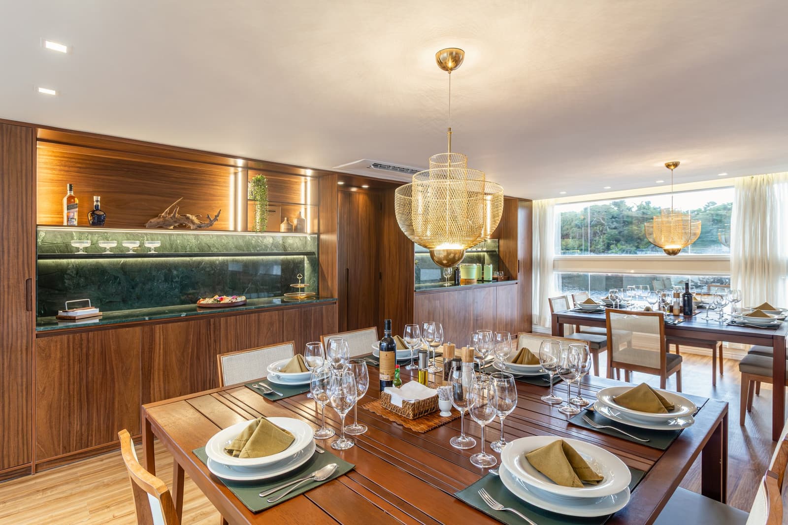 Good food tastes even better when the Amazon is drifting past the window. Photo: STUDIO CREATE Elegantly set dining room aboard a luxury Amazon cruise in Brazil, featuring two wooden tables laid with white plates, olive linen napkins, and multiple wine glasses, a backlit walnut bar with green marble countertop, and large windows overlooking the river