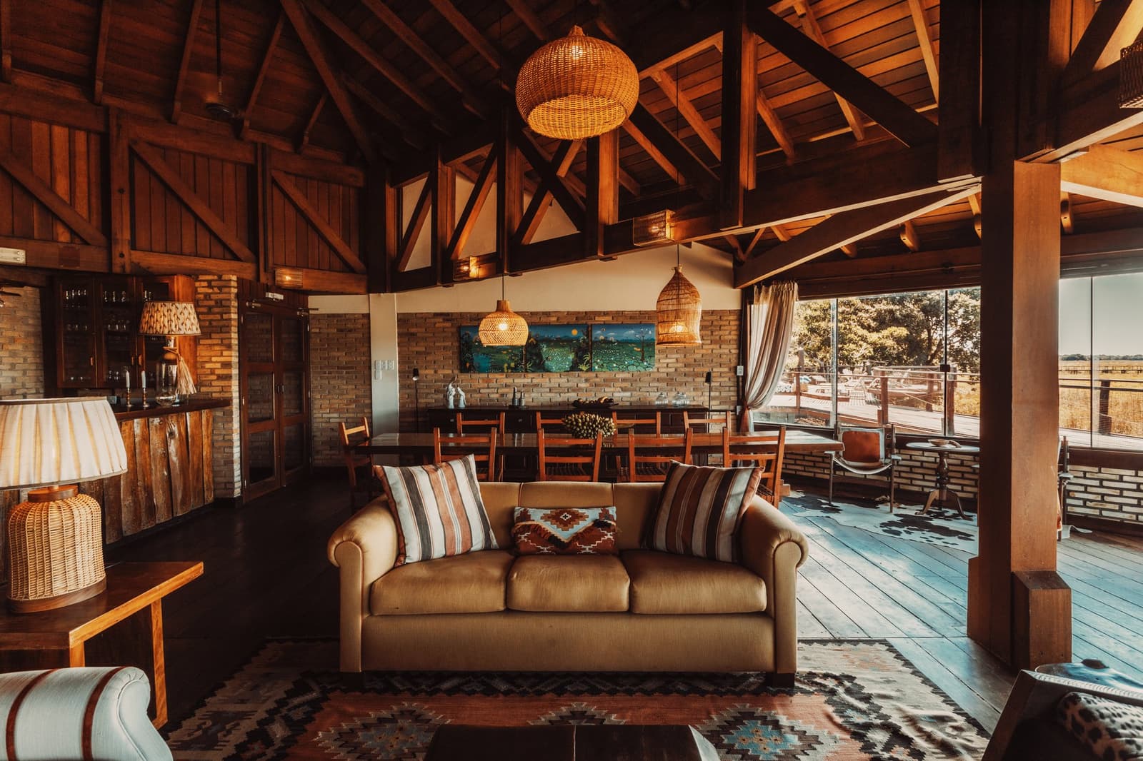 Sundowners taste better when the wetlands are your backdrop. Photo: Felipe Castellari Open-plan lodge lounge on a luxury Pantanal and Amazon trip, featuring a vaulted wooden ceiling with wicker pendant lights, a tan leather sofa with patterned cushions, a kilim rug, a dining area, and floor-to-ceiling windows opening onto a deck with savannah views beyond