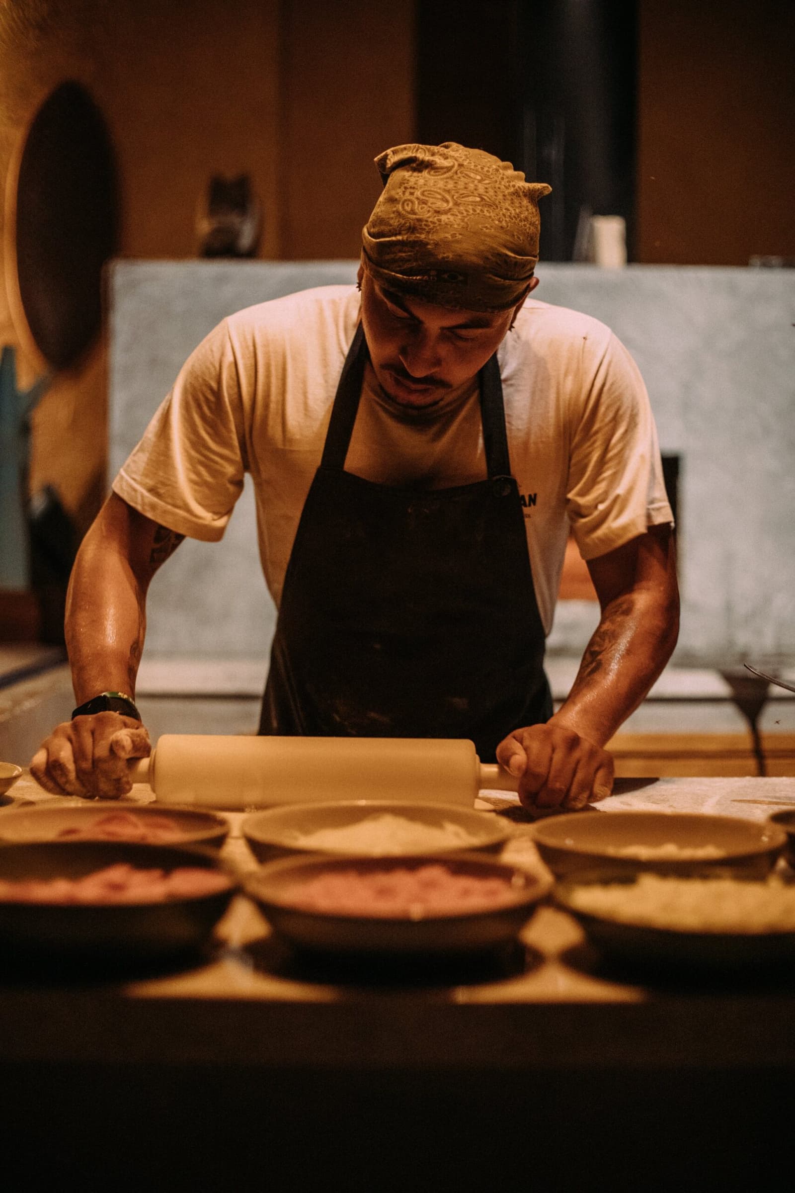 Hands deep in the dough, long before anyone sits down to eat. A tattooed chef wearing a bandana and dark apron rolls out pastry dough on a wooden counter, with several ceramic bowls of filling arranged in front of him, during preparation for a luxury Pantanal and Amazon trip lodge dinner
