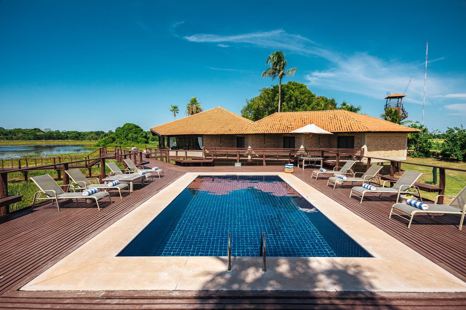 Pool time between safaris, not a bad way to spend a Pantanal afternoon. Photo: Felipe Castellari Rectangular swimming pool on a hardwood deck surrounded by sun loungers, with a tiled-roof lodge building and a wetland river view beyond, part of a luxury Pantanal and Amazon trip experience