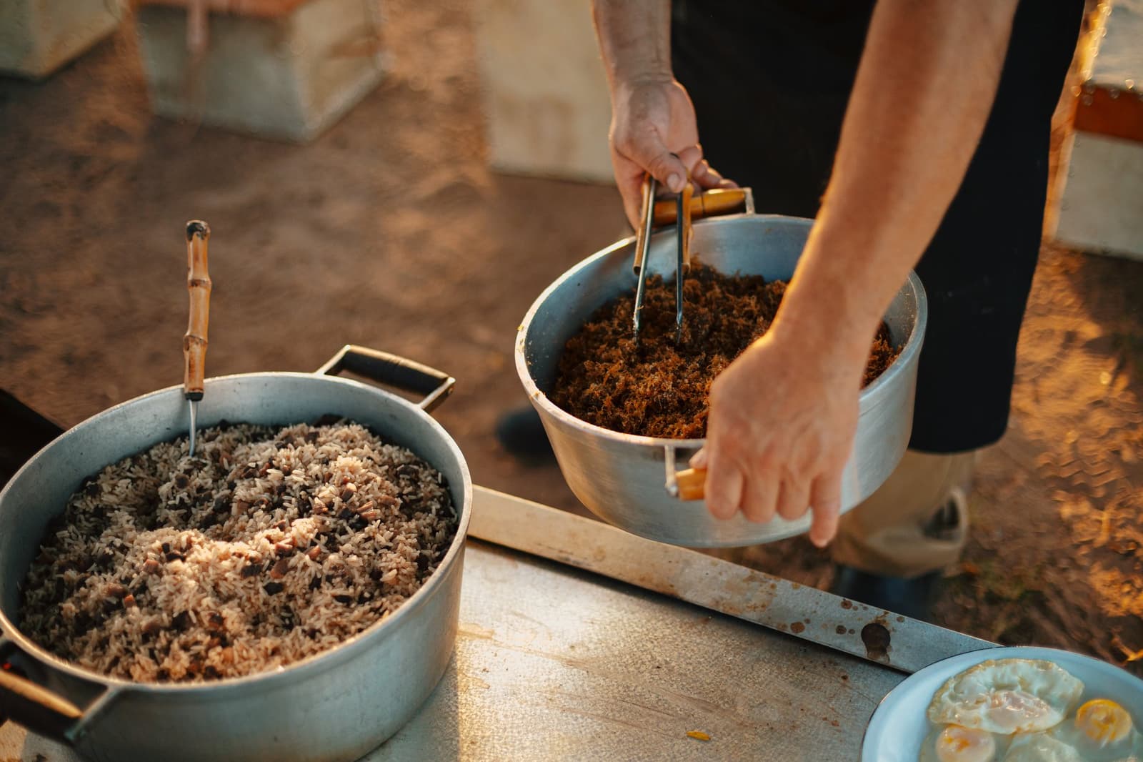 Rice, beans, shredded meat, fried eggs, and the kind of generosity that fills a table without fuss. A cook's hands stirring dark shredded meat in an aluminum pot beside a large pot of rice and black beans on an outdoor camp table, warm golden light catching the steam on this luxury Pantanal and Amazon trip