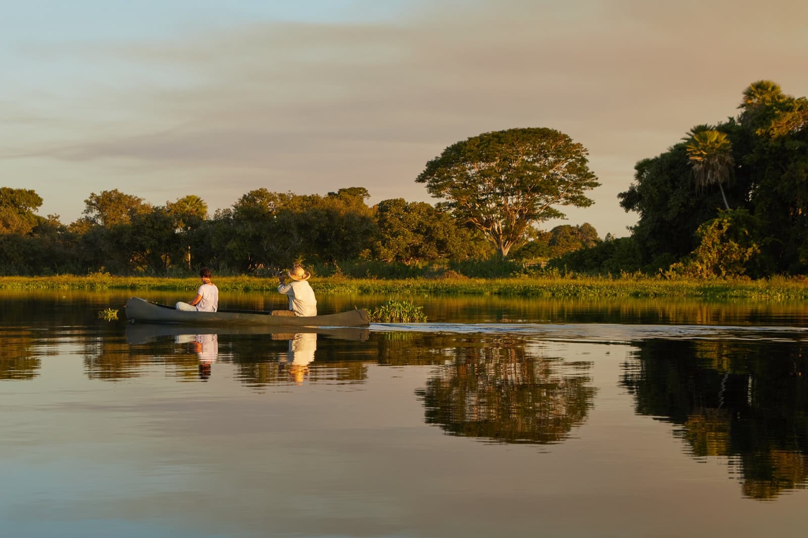 Paddling at dusk, the river holds the whole sky in its hands. Two travelers sit quietly in a canoe on a glassy wetland river at golden hour, reflected perfectly in the still water, with lush gallery forest lining the far bank — a defining moment of a luxury Pantanal and Amazon trip