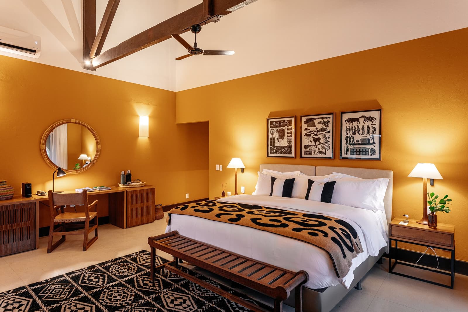 Folk art on the walls, a ceiling fan overhead, and a bed you'll have trouble leaving before the 5:30 AM safari. Photo: Felipe Castellari Warm ochre-walled guest room on a luxury Pantanal and Amazon trip, featuring a king bed with a patterned throw, exposed wooden ceiling beams, a ceiling fan, three Brazilian folk-art prints above the headboard, and a rattan-framed round mirror above a wooden desk